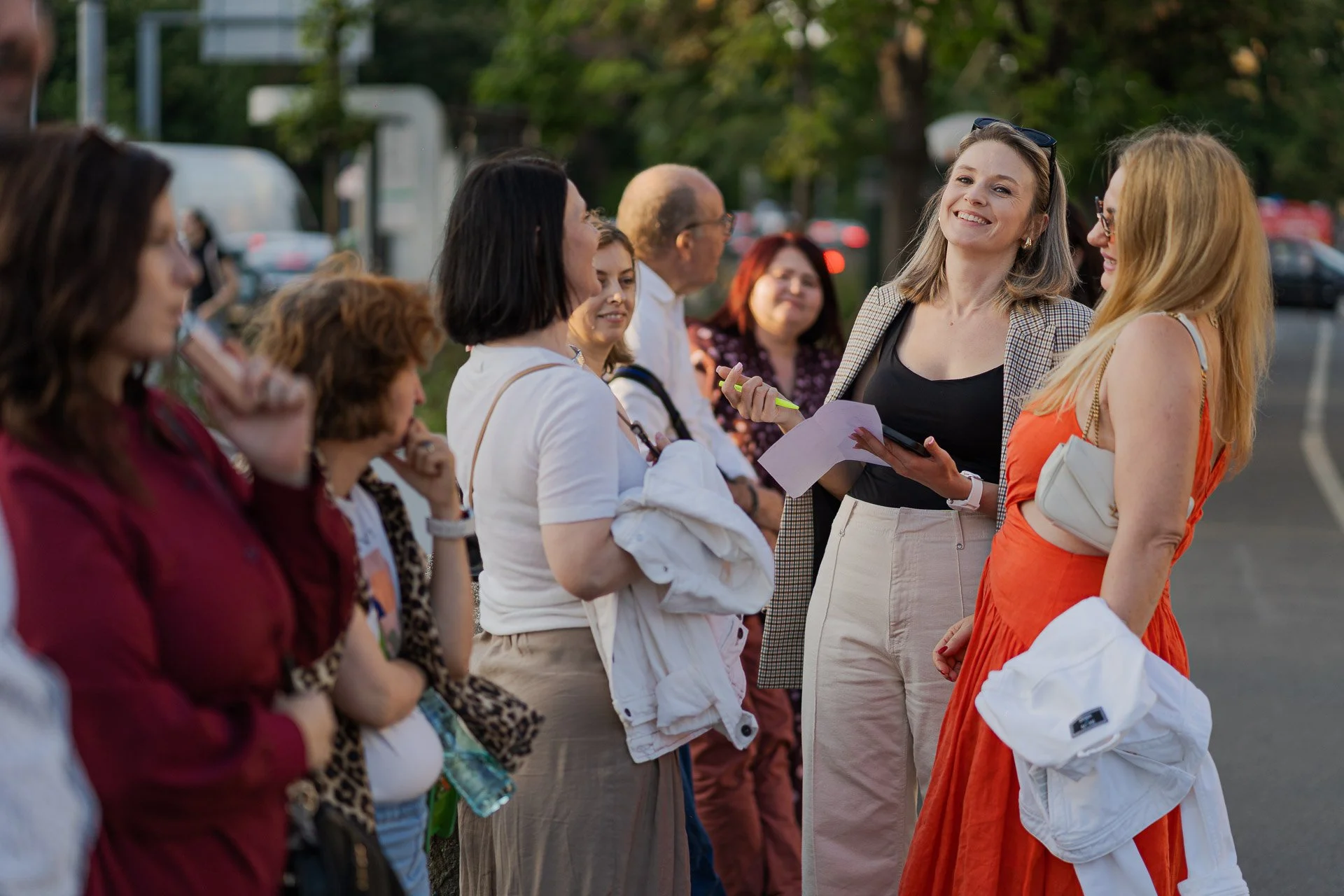 A group of women are standing outdoors, engaged in conversation, with one woman smiling and holding a notepad and pen, wearing sunglasses on her head, while others listen and interact.