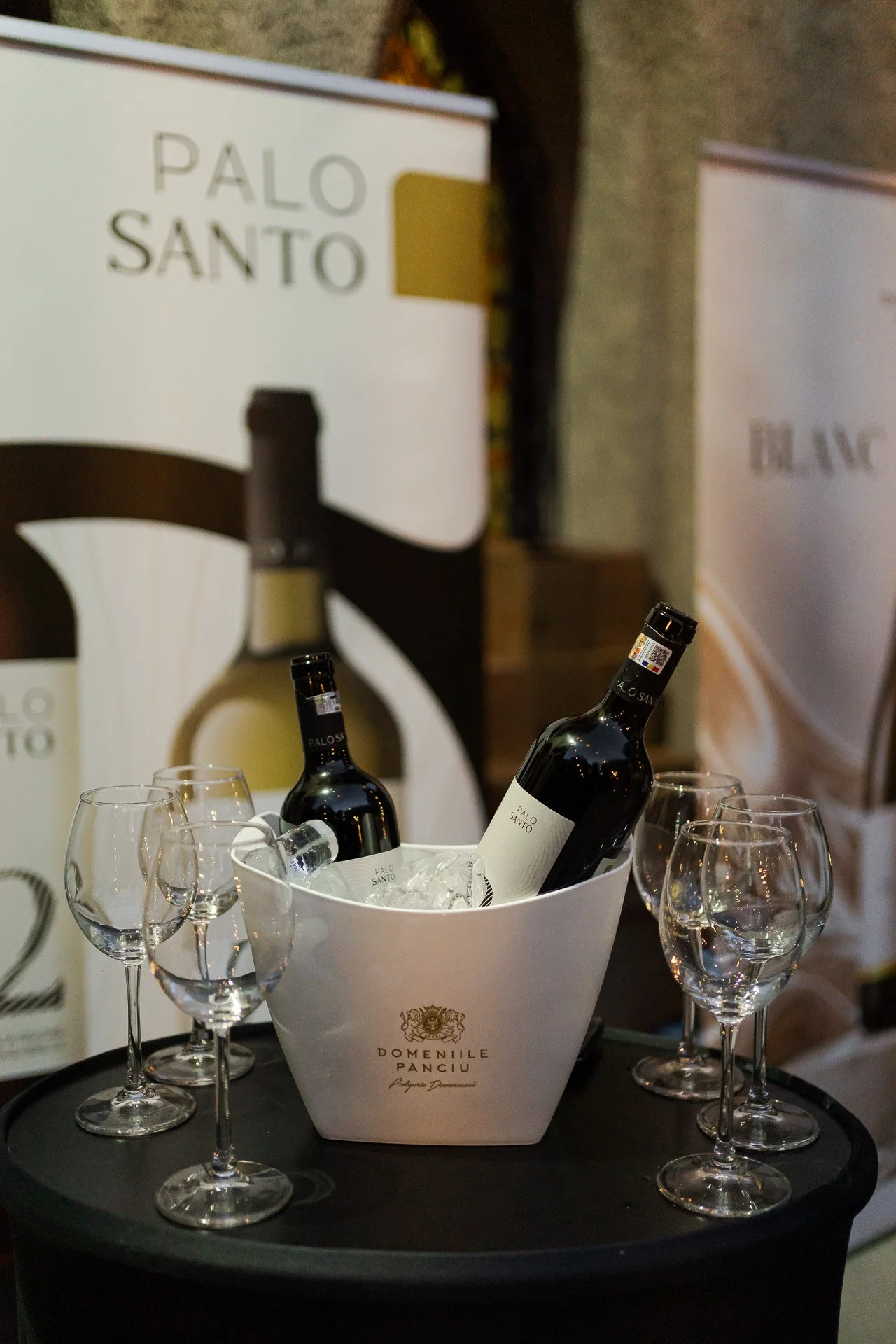 Several wine glasses surrounding a white bucket with two bottles of wine in ice, on a black tray. The bucket has the logo and name DOMEÑILE PANCIU and is on a black surface. In the background, there are posters or signs, one with the label PALO SANTO