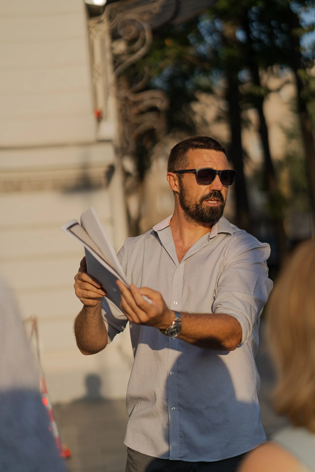 A man with sunglasses, a beard, and a watch is holding and showing a newspaper to a woman, outdoors on a sunny day, with trees in the background.