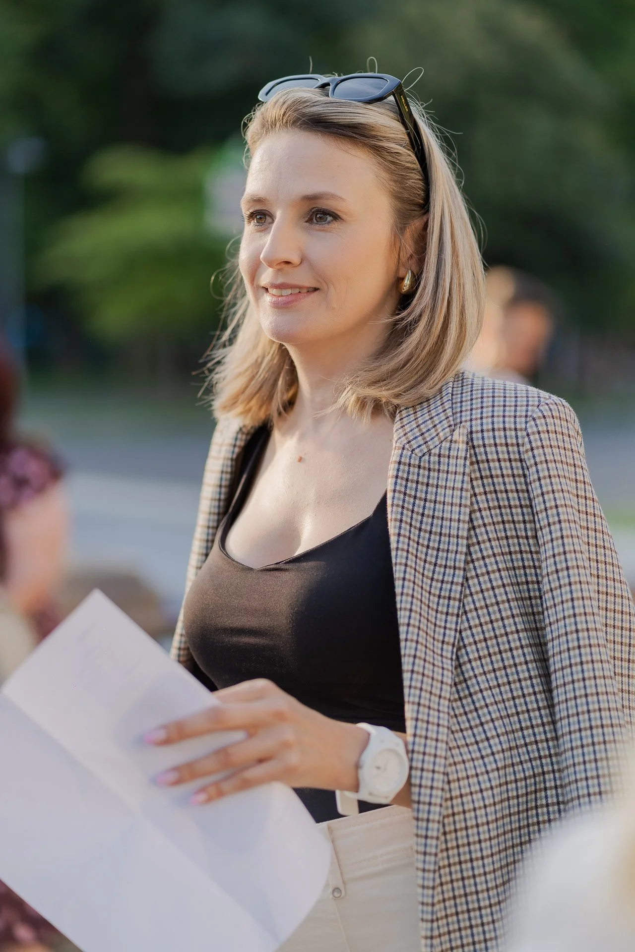 A woman with blonde hair, wearing sunglasses on her head, a plaid blazer, a black top, earrings, and a white wristwatch, is holding and looking at a white piece of paper outdoors.