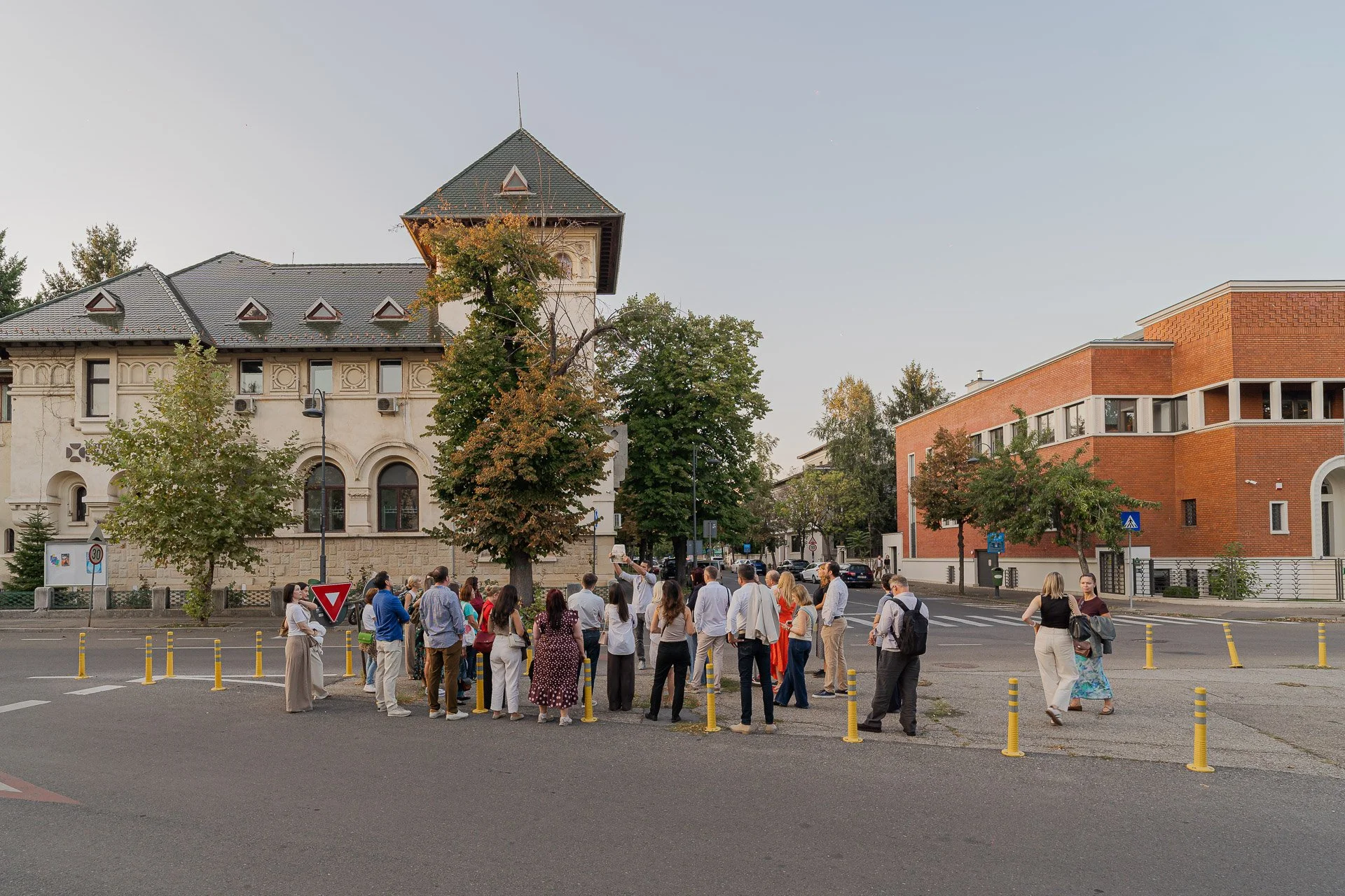 A group of people waiting to cross the street at a pedestrian crosswalk, with trees and buildings in the background.