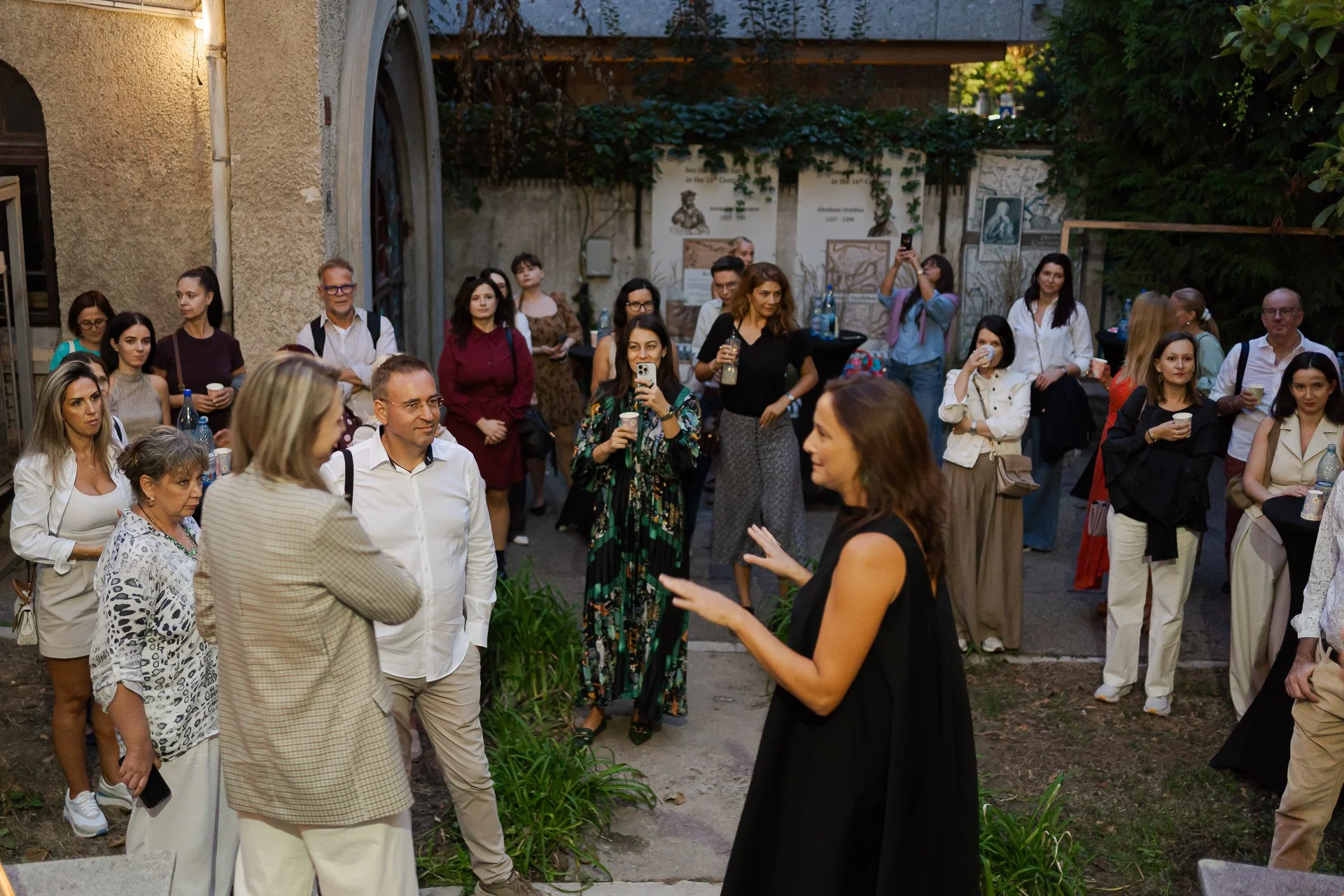 A group of people gathered outdoors, engaged in conversation and listening to a woman speaking. Several attendees are holding drinks or cups, and some are taking photos or videos. The scene appears to be a social or professional event held outside ne