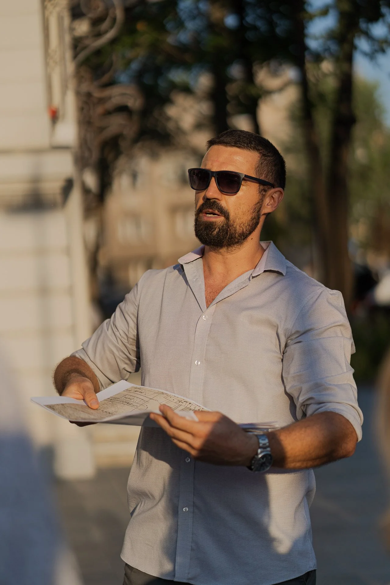 A man wearing sunglasses and a light-colored shirt, holding papers or a brochure, outdoors on a sunny day.