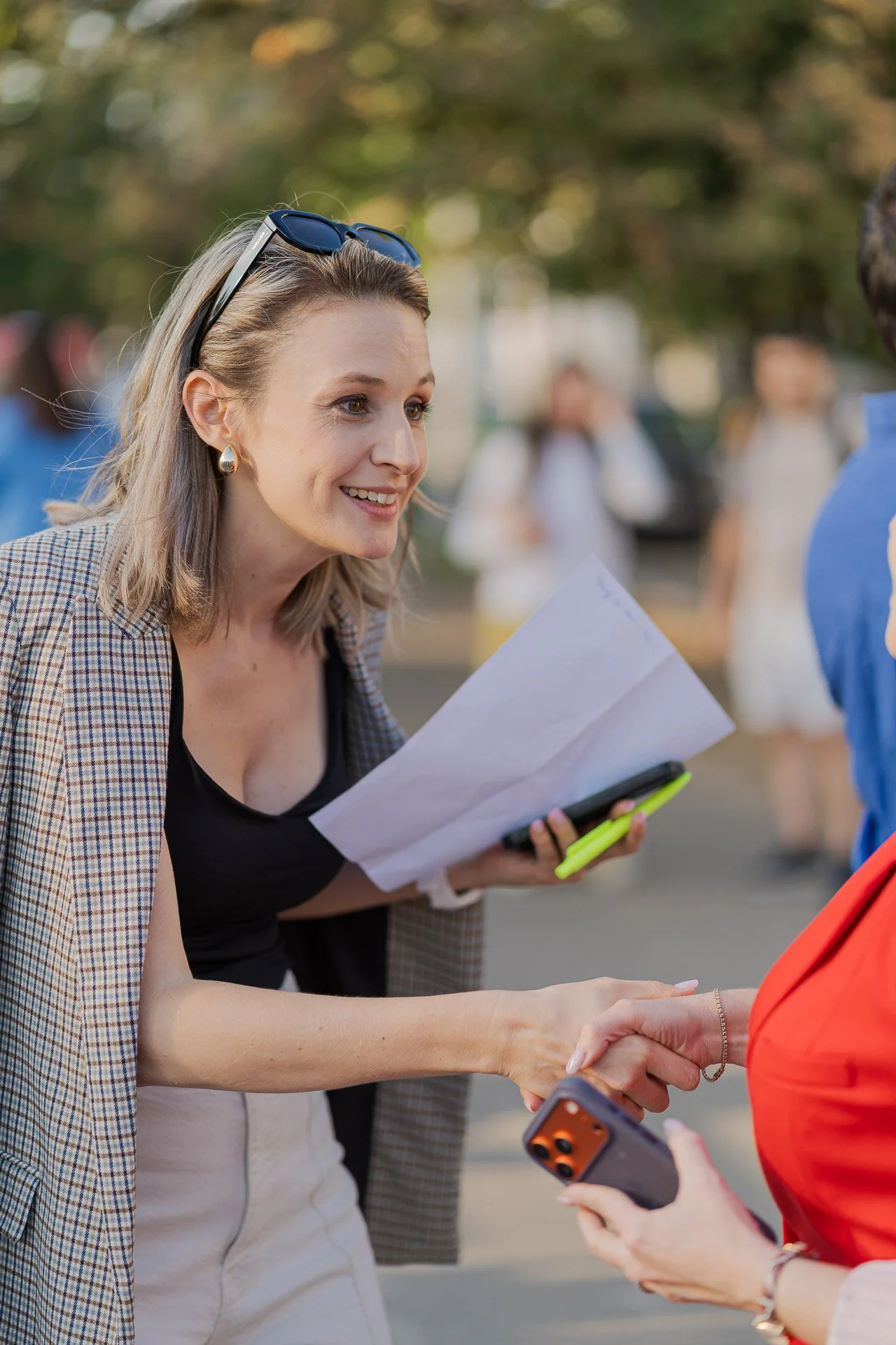 A woman with blonde hair, wearing sunglasses on her head, a checked blazer, and a black top, is smiling and shaking hands with another person in a red jacket. She is holding a paper and a phone with a yellow case.