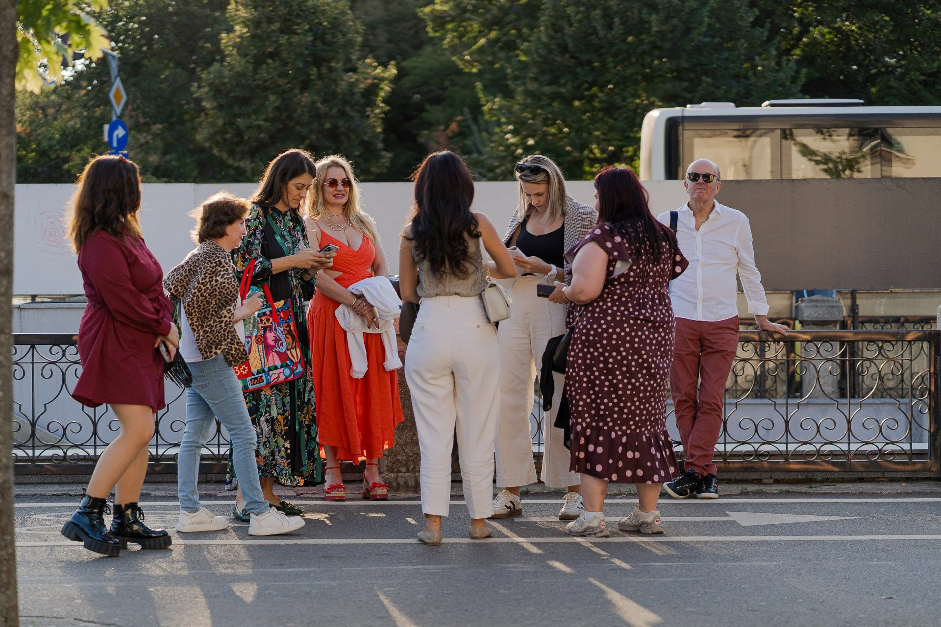 Group of people standing outdoors behind a decorative railing, engaged in conversation, with trees and a bus in the background.
