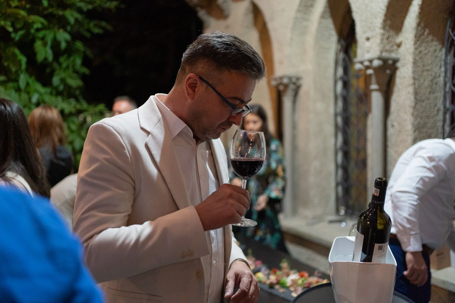 A man in a white suit holding a glass of red wine at a social gathering. The background shows other people and a stone wall with decorative arches.