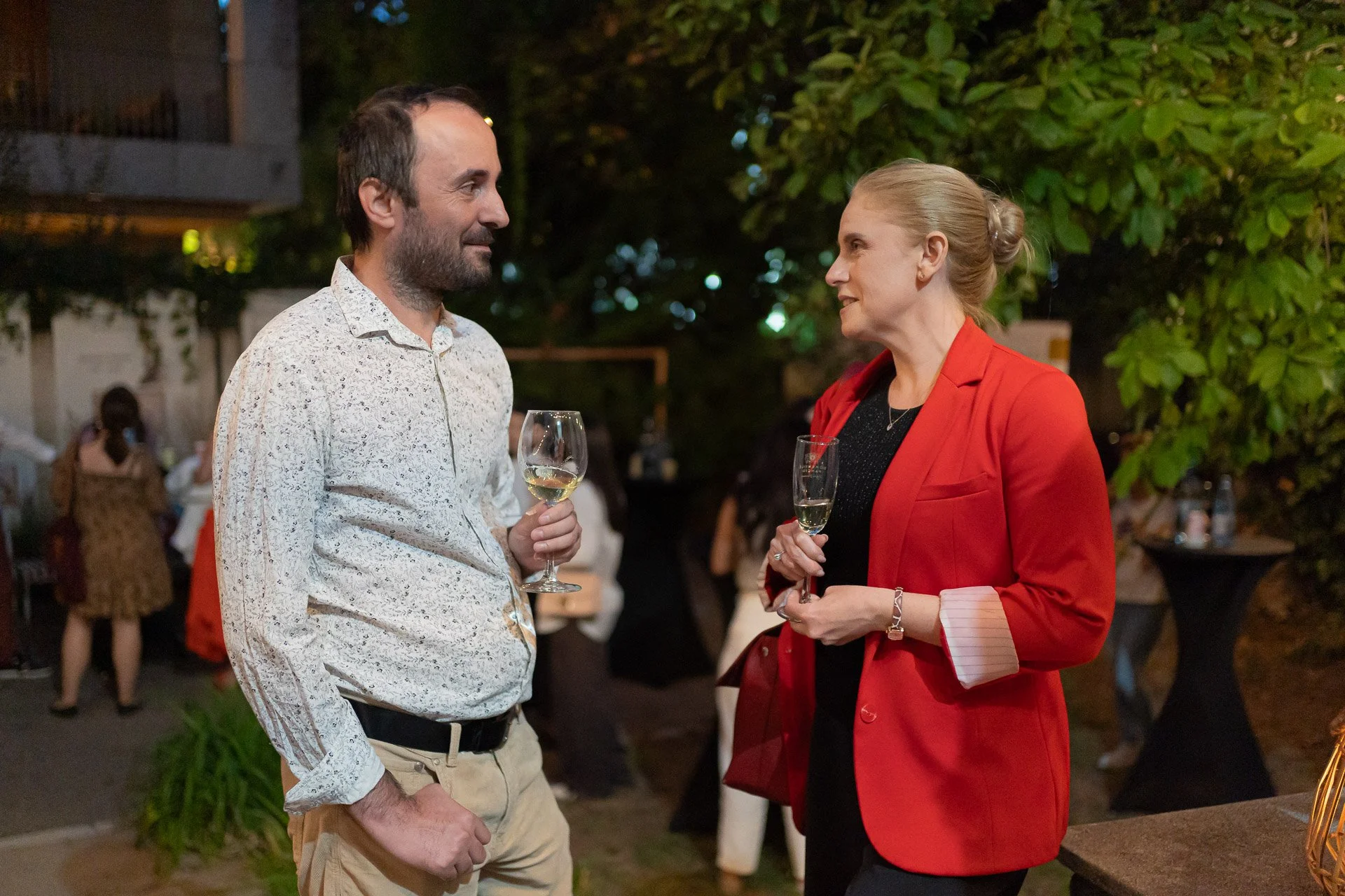 A man and woman are standing outdoors at night, engaged in conversation while holding wine glasses. The man has short dark hair and a beard, wearing a patterned white shirt. The woman has blonde hair pulled back, wearing a red blazer over a black top