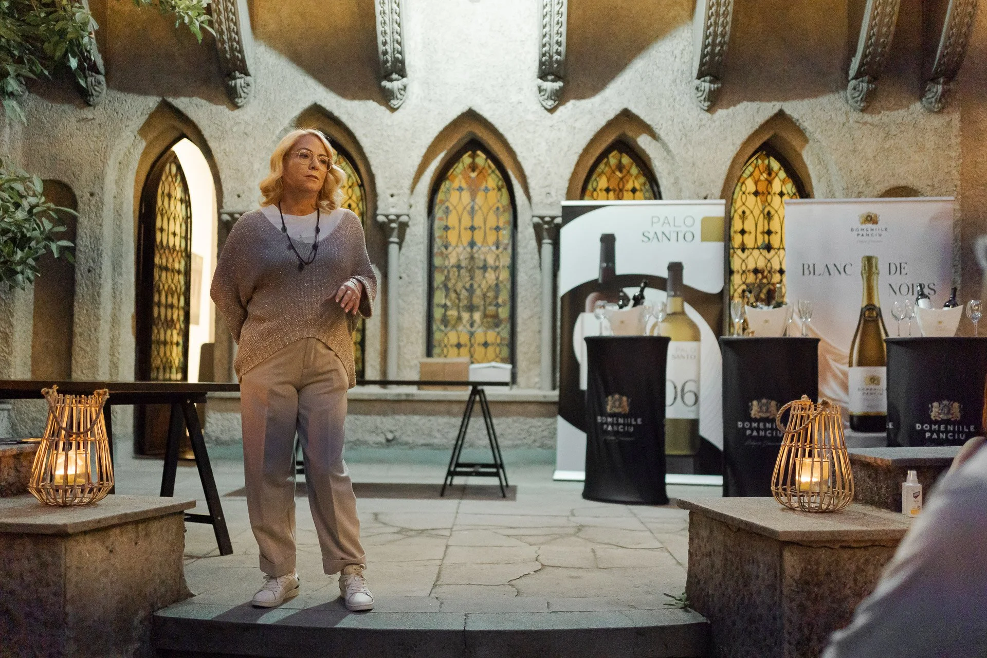 A woman with glasses, blonde hair, wearing a beige sweater, pants, and white sneakers, is standing on a small platform in an indoor event space. The background features a stone wall with arched stained glass windows, banners with wine bottles, and wa
