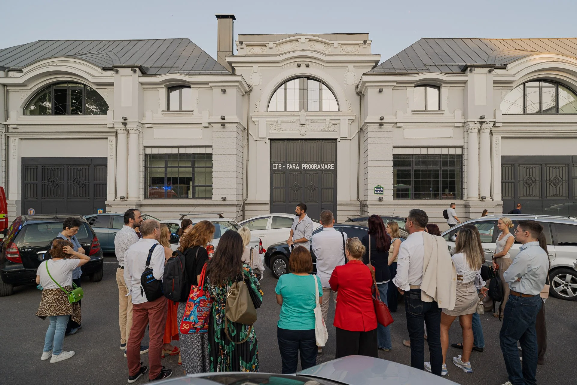 A large group of people gathered in front of a white historic building with arched windows and a sign that reads 'I.T.P. - Fara Programare.' Some people are talking, walking, or standing around cars parked along the street.