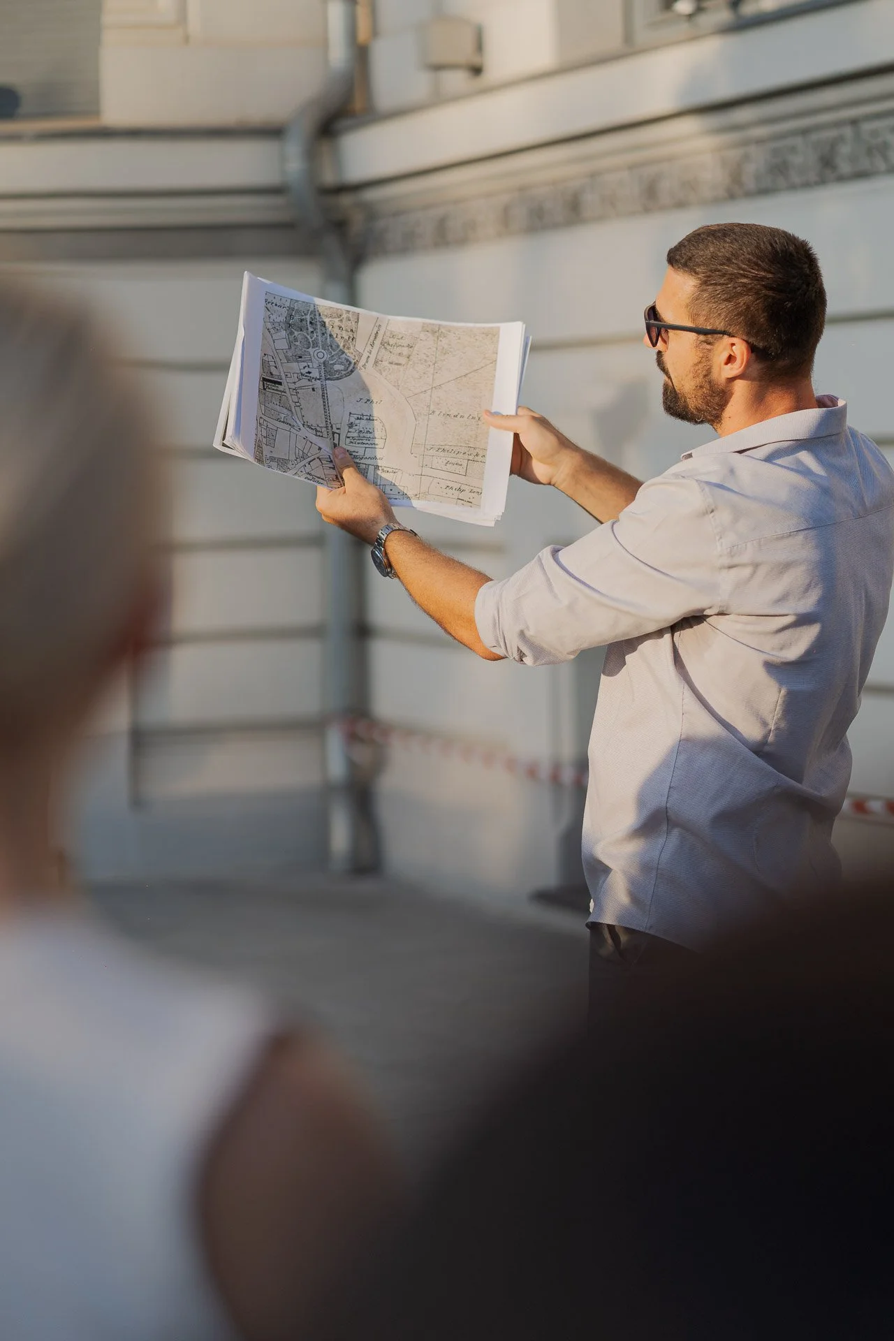 A man wearing sunglasses and a light-colored shirt holding and examining an architectural map or blueprint outdoors near a building.