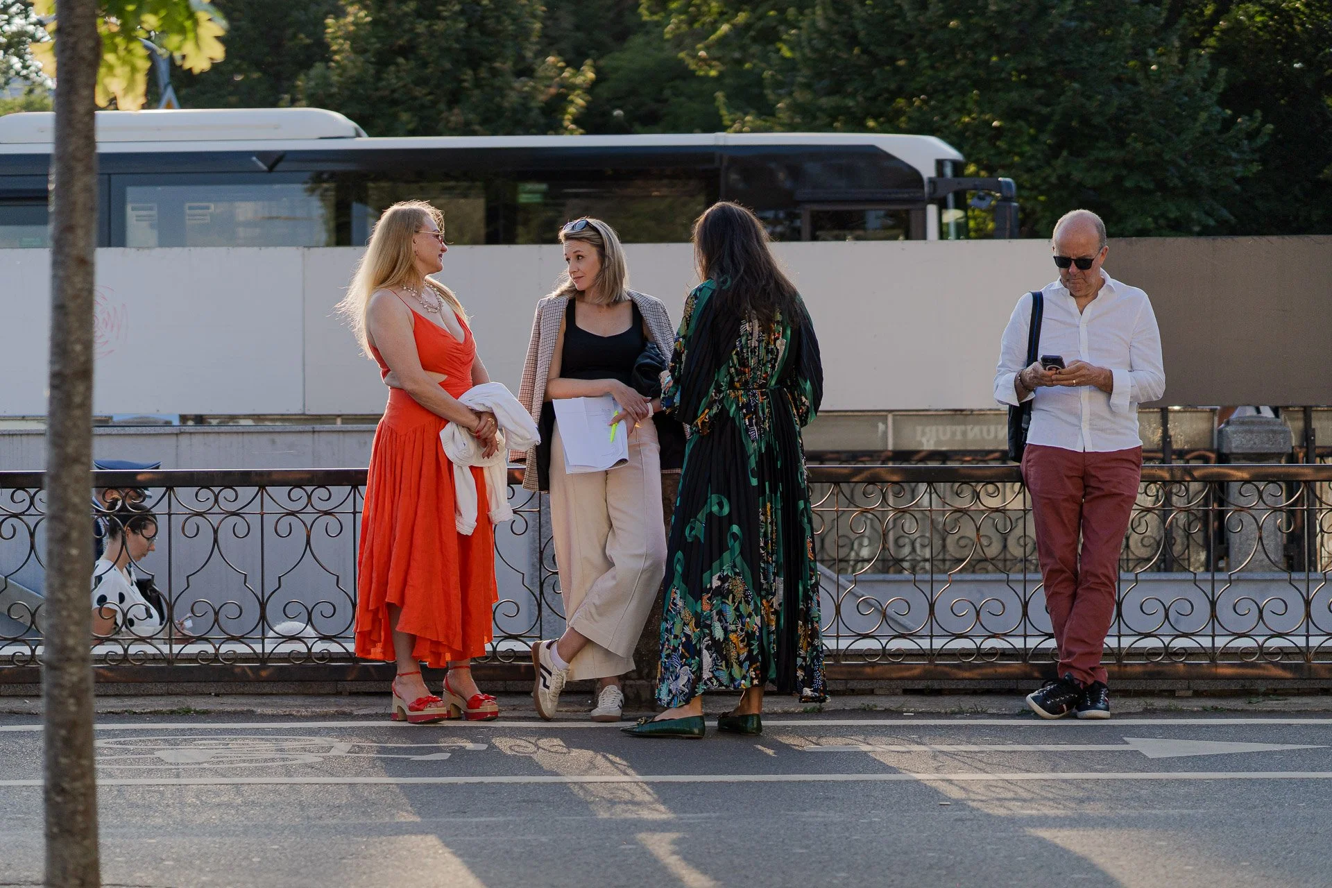Four women and one man standing and talking on a sidewalk in front of a white fence and a bus in the background. The women are dressed in colorful outfits, and the man is using a cellphone.
