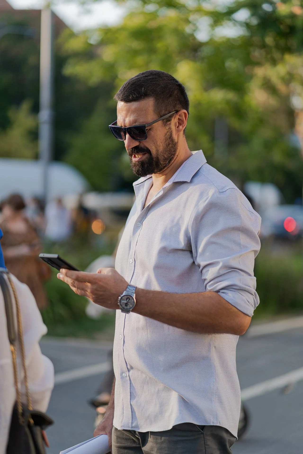 A man wearing sunglasses and a light-colored button-up shirt looking at his phone outdoors, with blurred trees and people in the background.