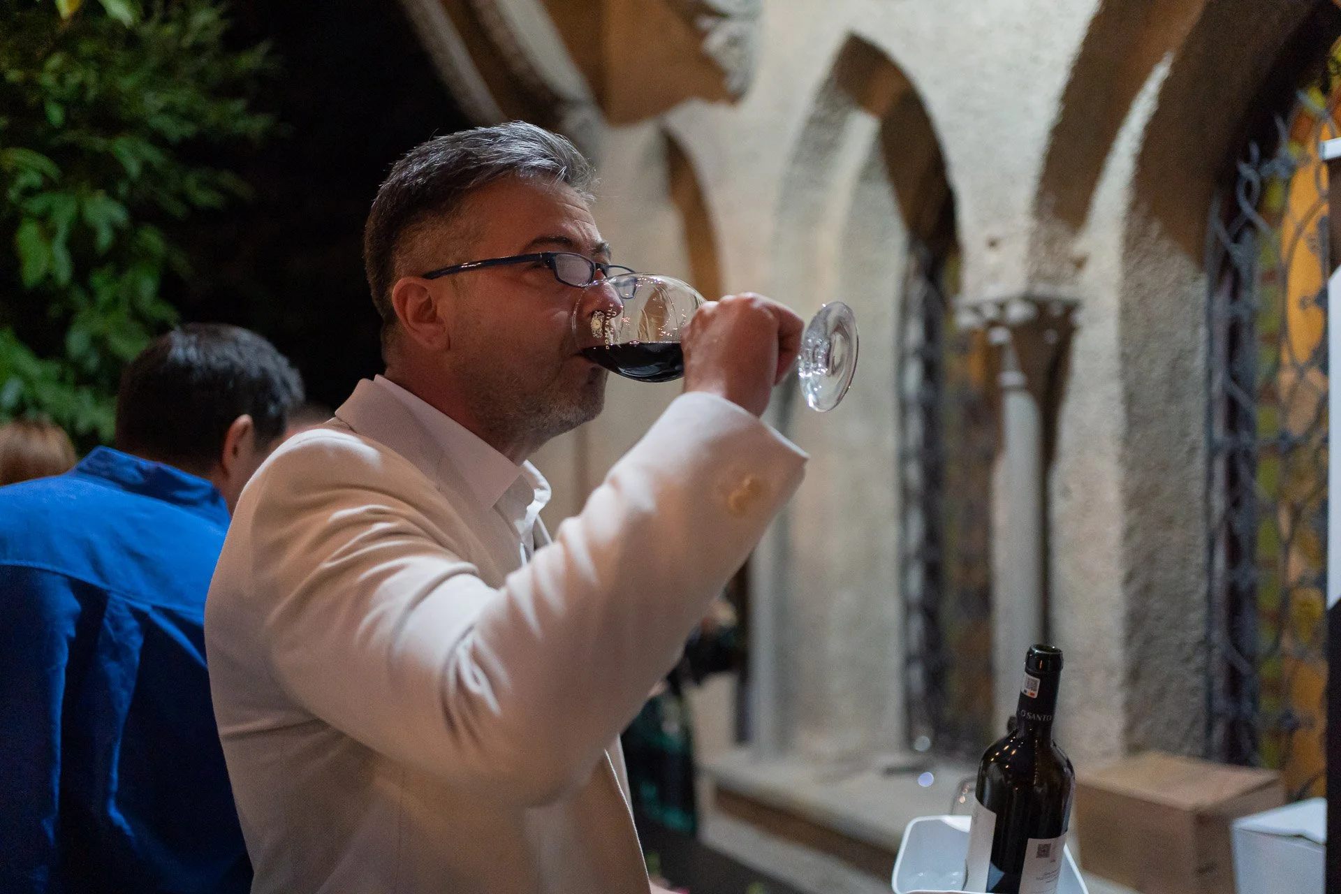 A man with gray hair and glasses wearing a white blazer is drinking red wine from a glass at an indoor gathering. He is standing near a wine bottle on a table, with arched stone walls and greenery in the background.