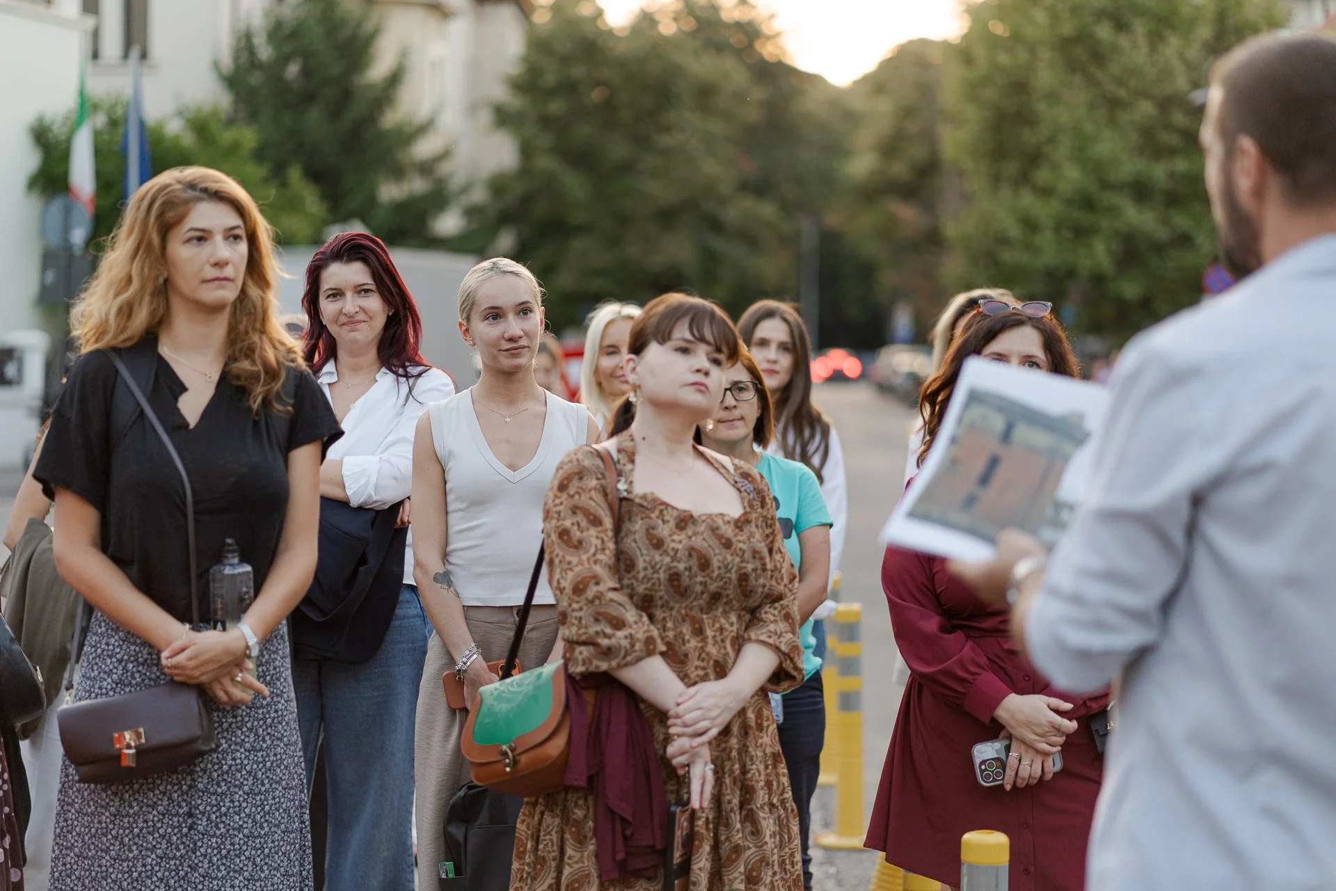 A group of diverse women listening attentively to a man giving a presentation on a city street during early evening.