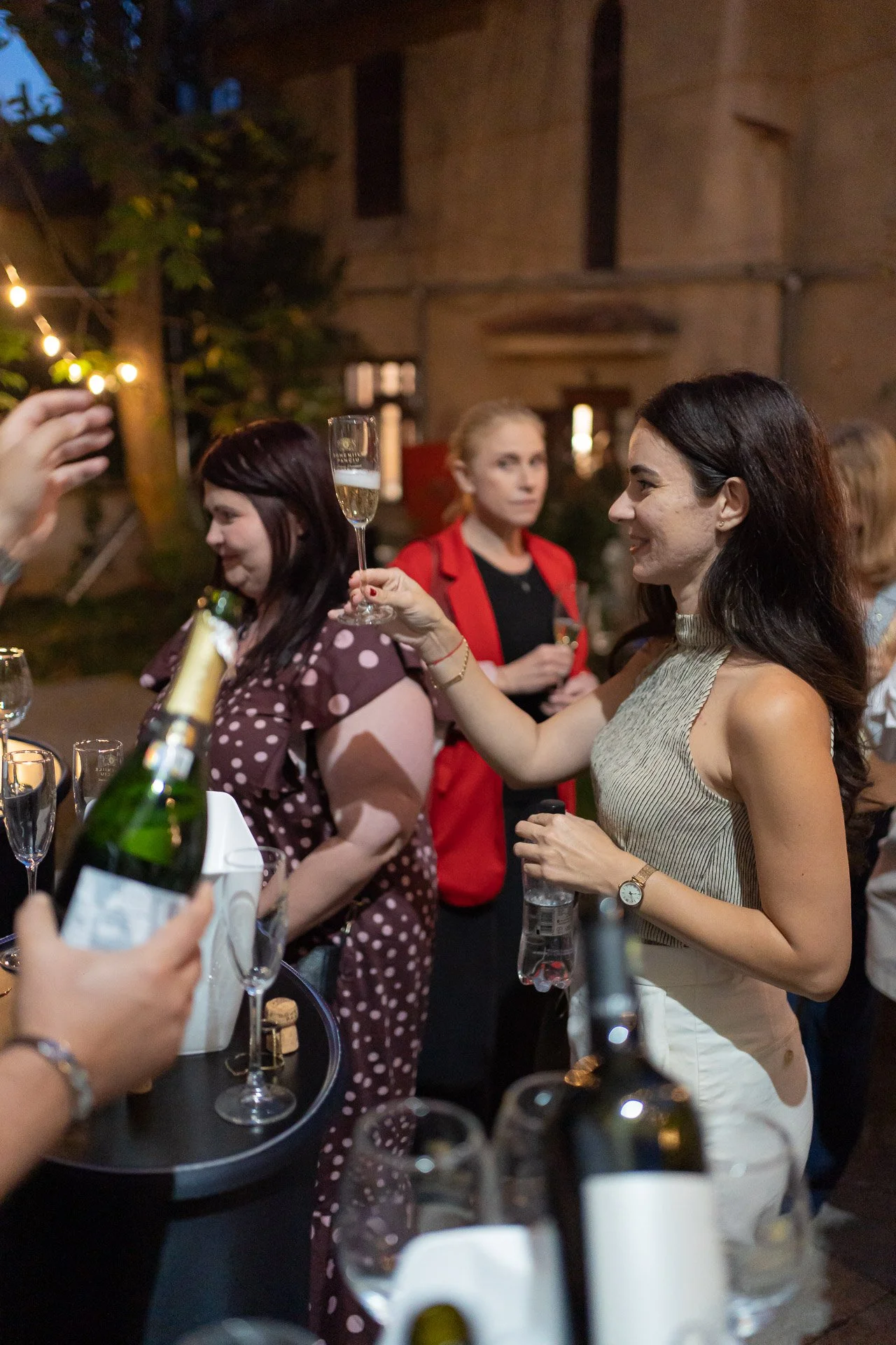 Women at a social gathering with drinks, including champagne, in a cozy, dimly lit indoor setting with string lights and greenery.