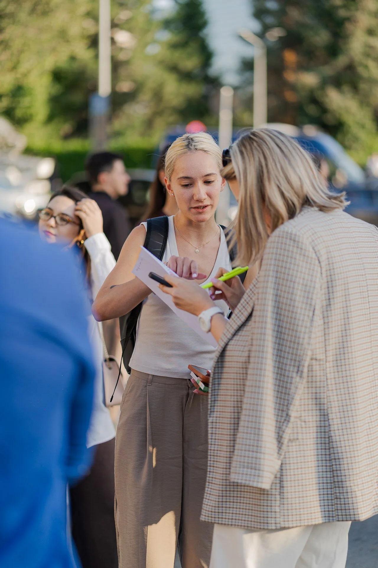 Two women are looking at a phone and a piece of paper together outdoors, with several people in the background and green trees.