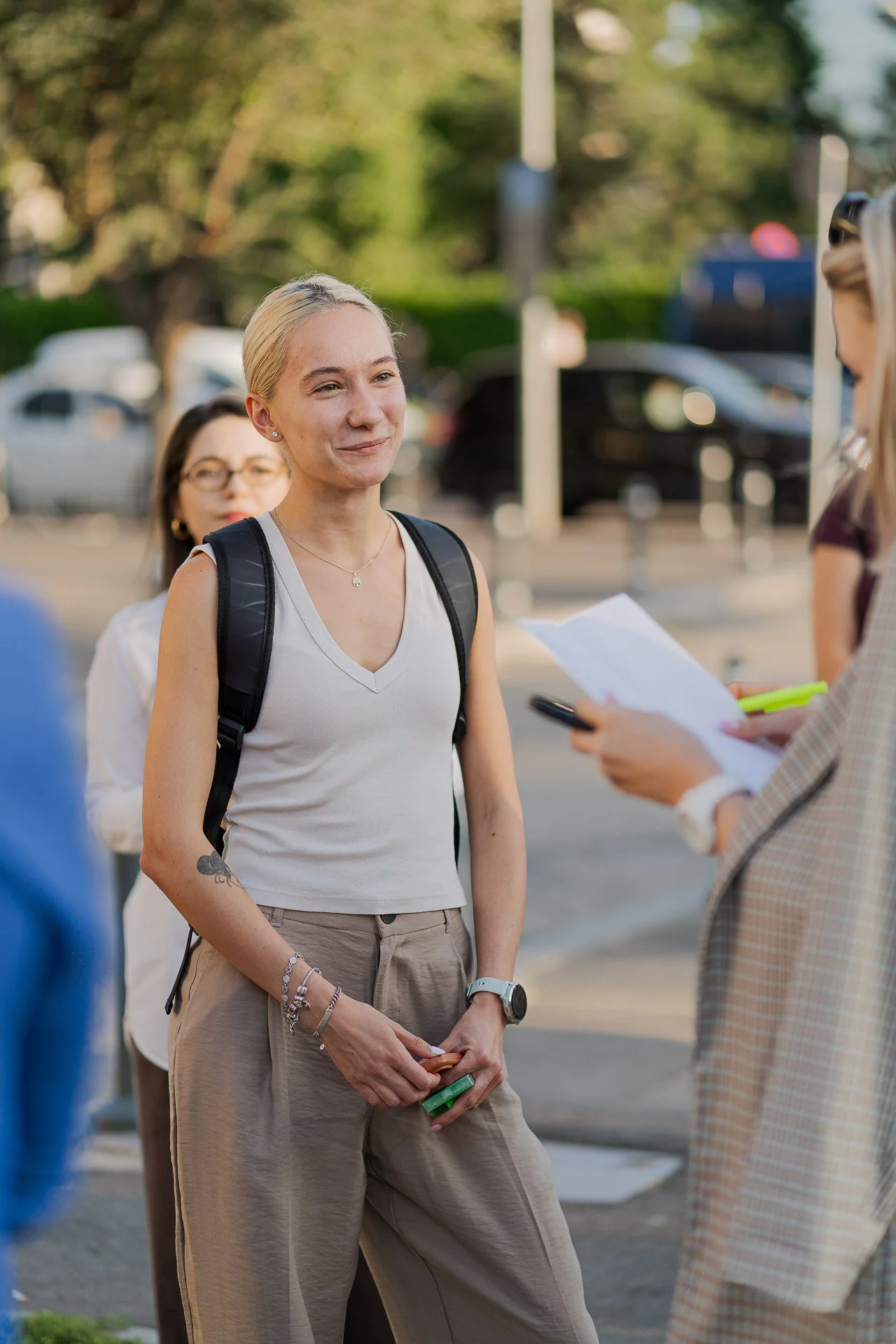 A young woman with blonde hair, jewelry, and a backpack smiling outdoors while talking to another person holding papers and a pen.