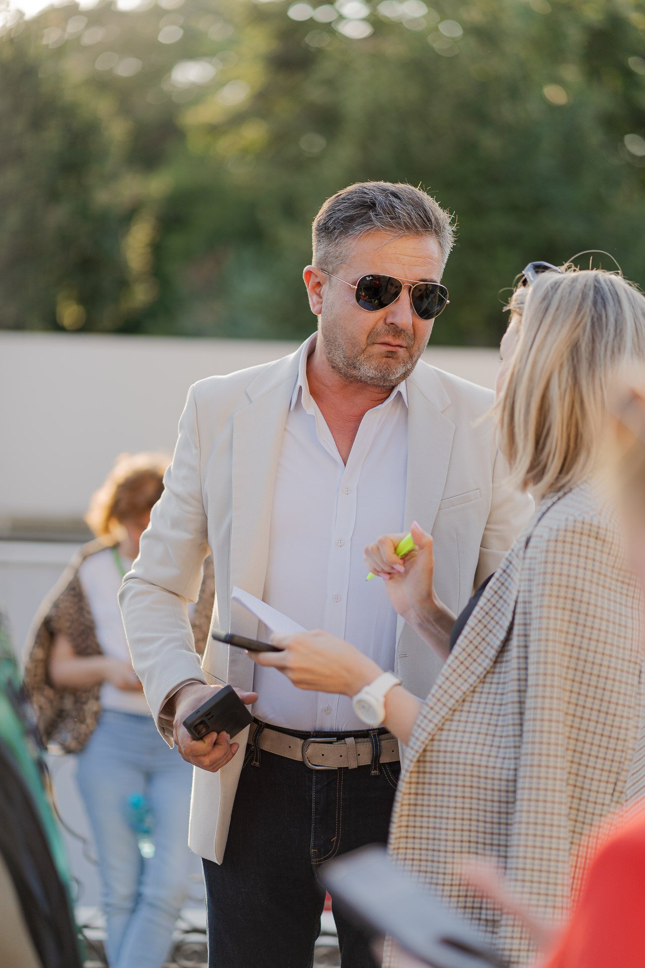 A man wearing a white blazer and sunglasses is engaged in conversation with a woman holding a paper and highlighter. They are outdoors with trees in the background and other people nearby.