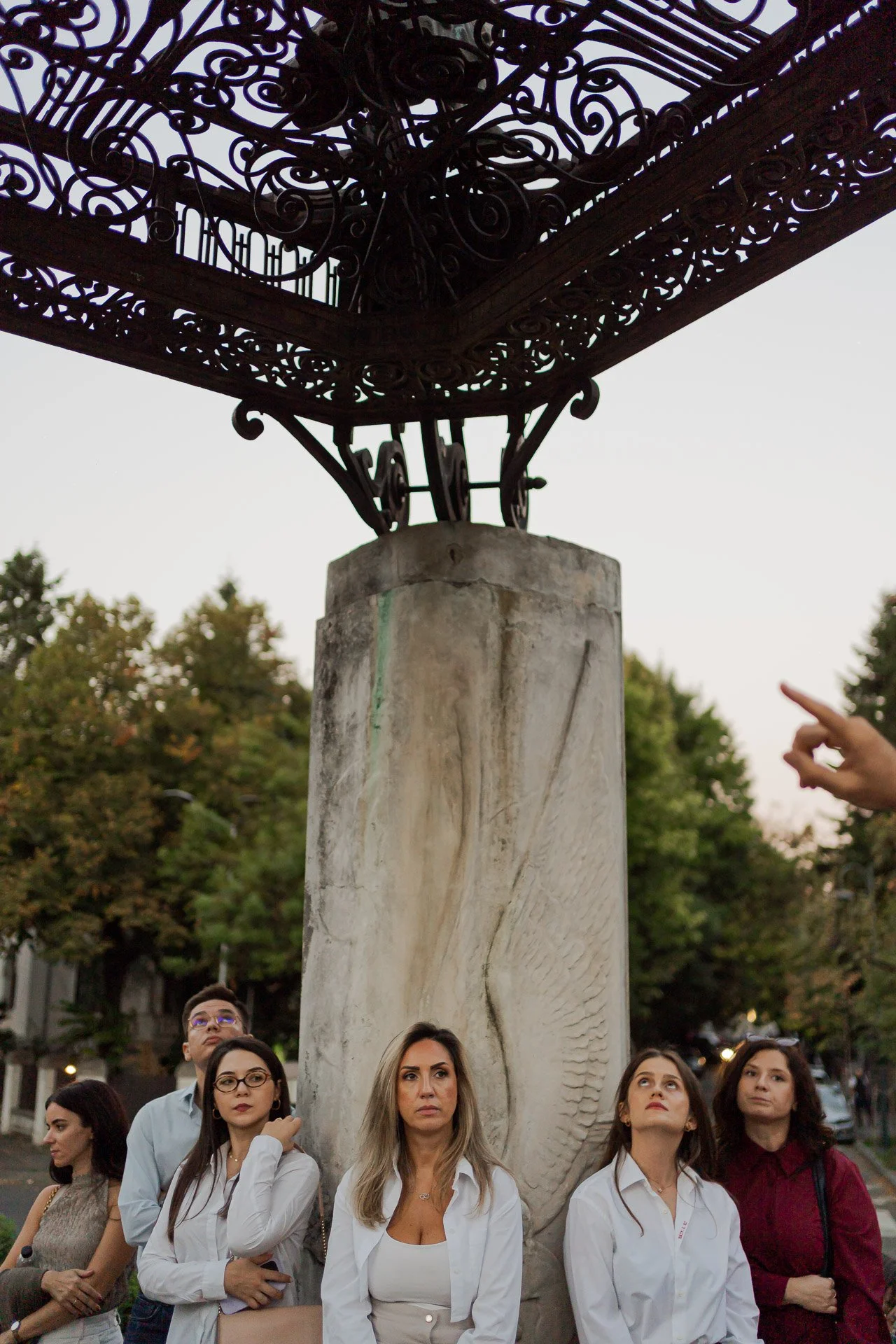 Group of women and men standing outdoors in front of a tall stone structure with intricate wrought iron detailing above, trees in the background, and an overcast sky.