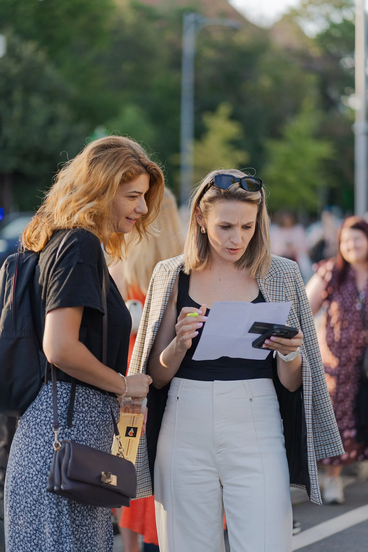Two women standing together outdoors, looking at a piece of paper and a smartphone. One woman has red hair and the other has blonde hair with sunglasses on her head.