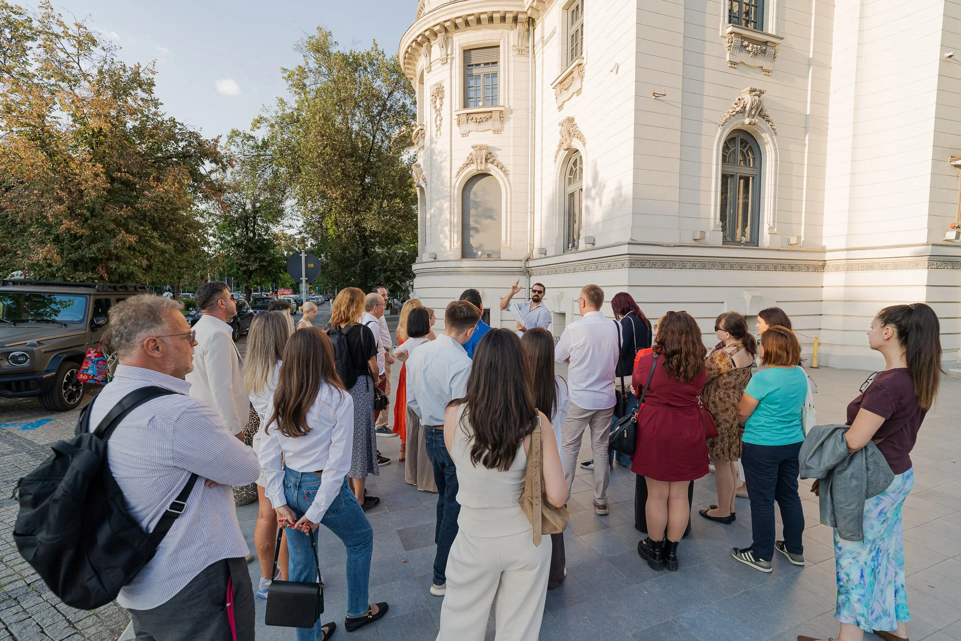 A diverse group of people gathered outdoors in front of a historic building with ornate white facade, listening to a tour guide who is gesturing and explaining.