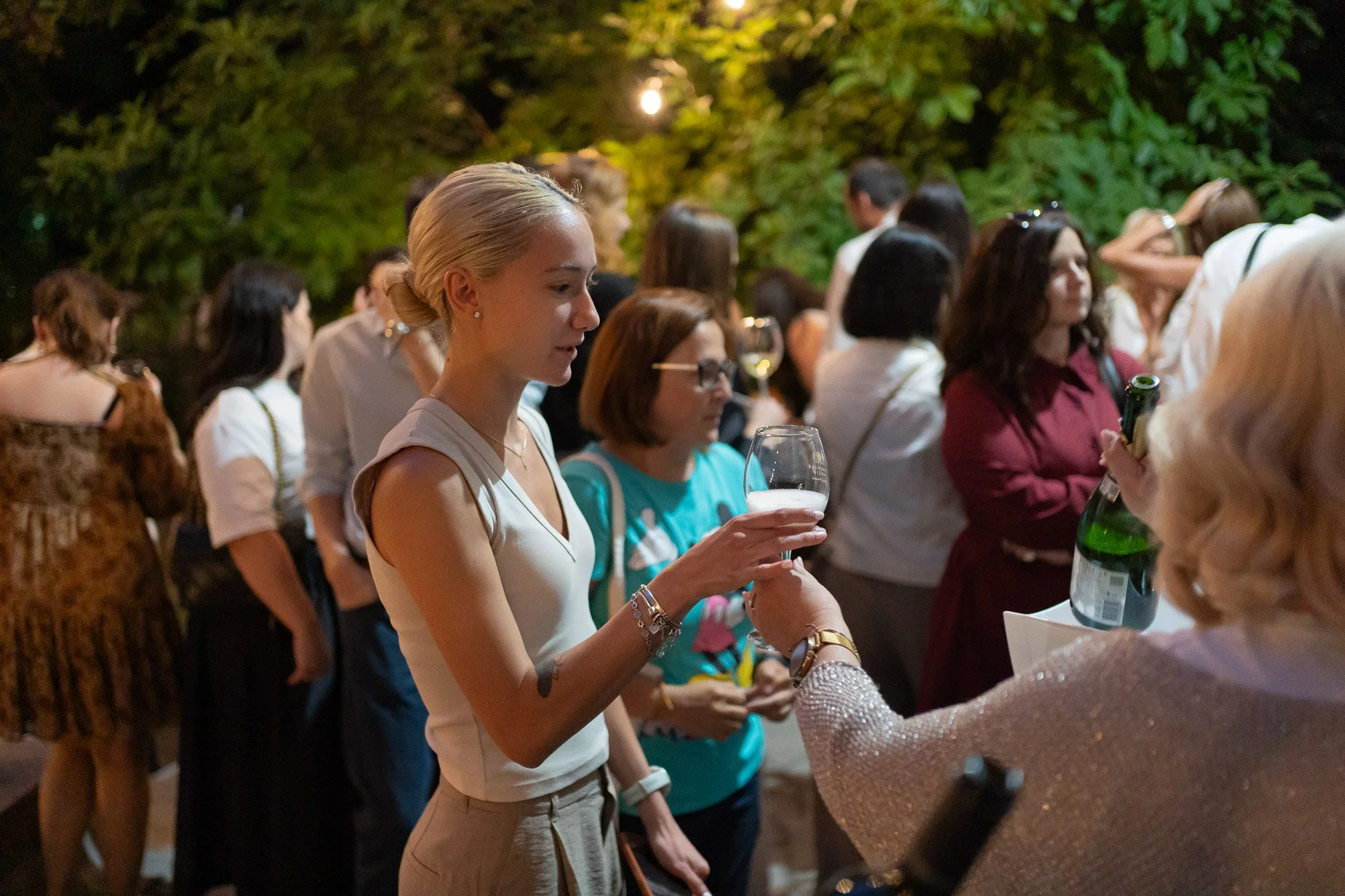 A woman with blonde hair styled in a low bun wearing a sleeveless cream blouse and jewelry, holding a wine glass, socializing at an outdoor gathering at night, with many people in the background surrounded by green foliage.