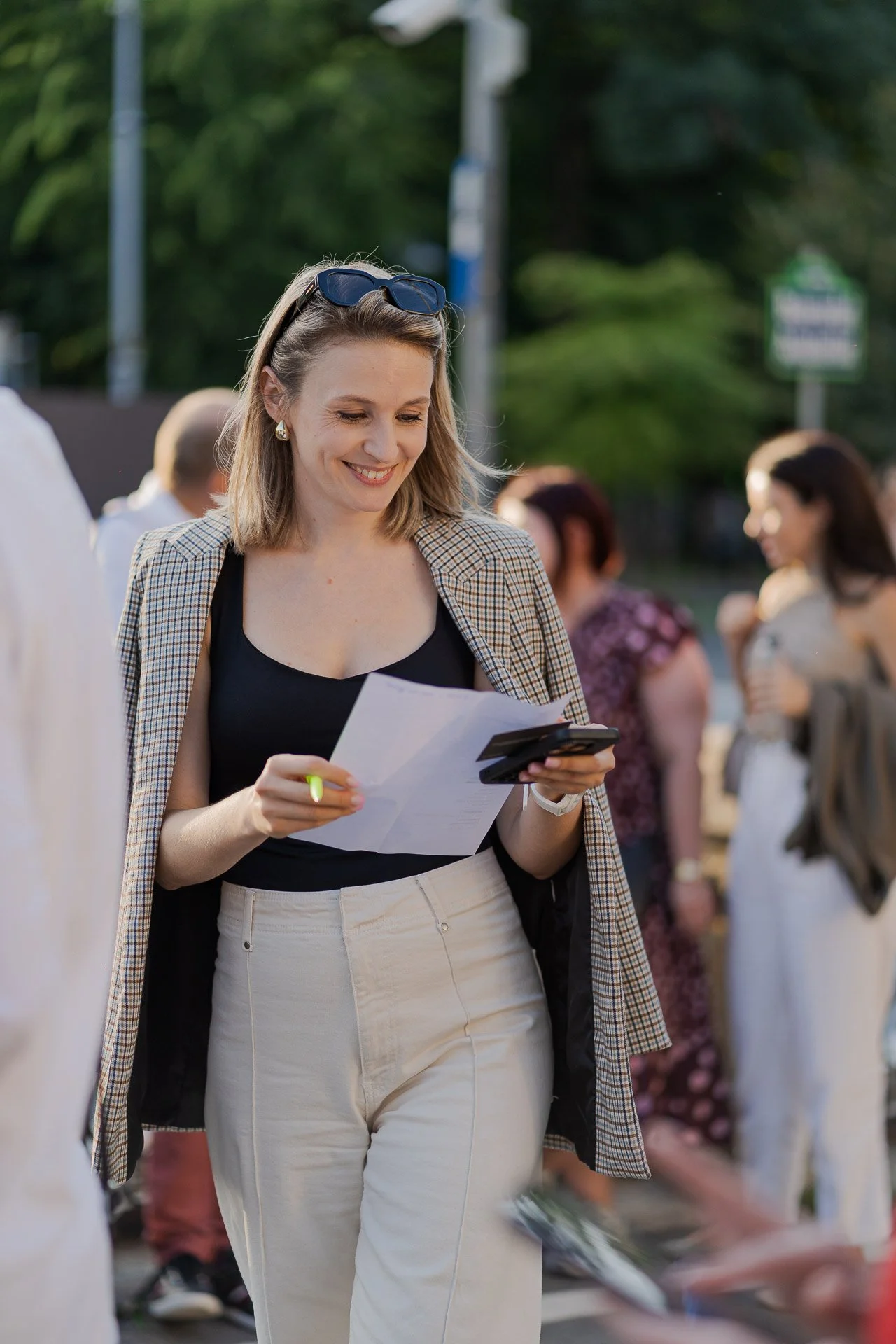 A woman smiling while looking at a piece of paper and a smartphone, outdoors with several people in the background.