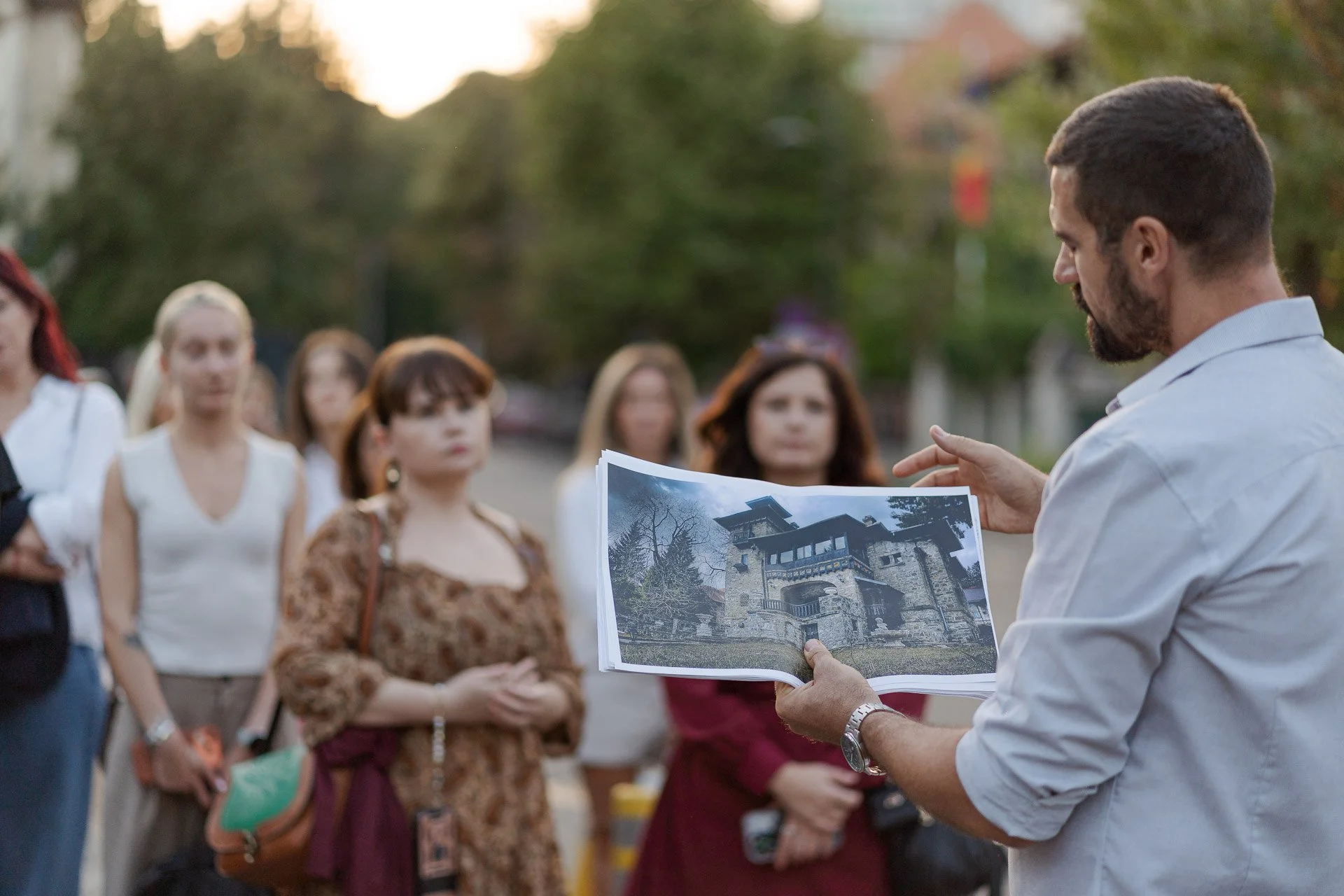 A man in a light-colored shirt showing a photo album to a group of women gathered outdoors during sunset, with trees in the background.