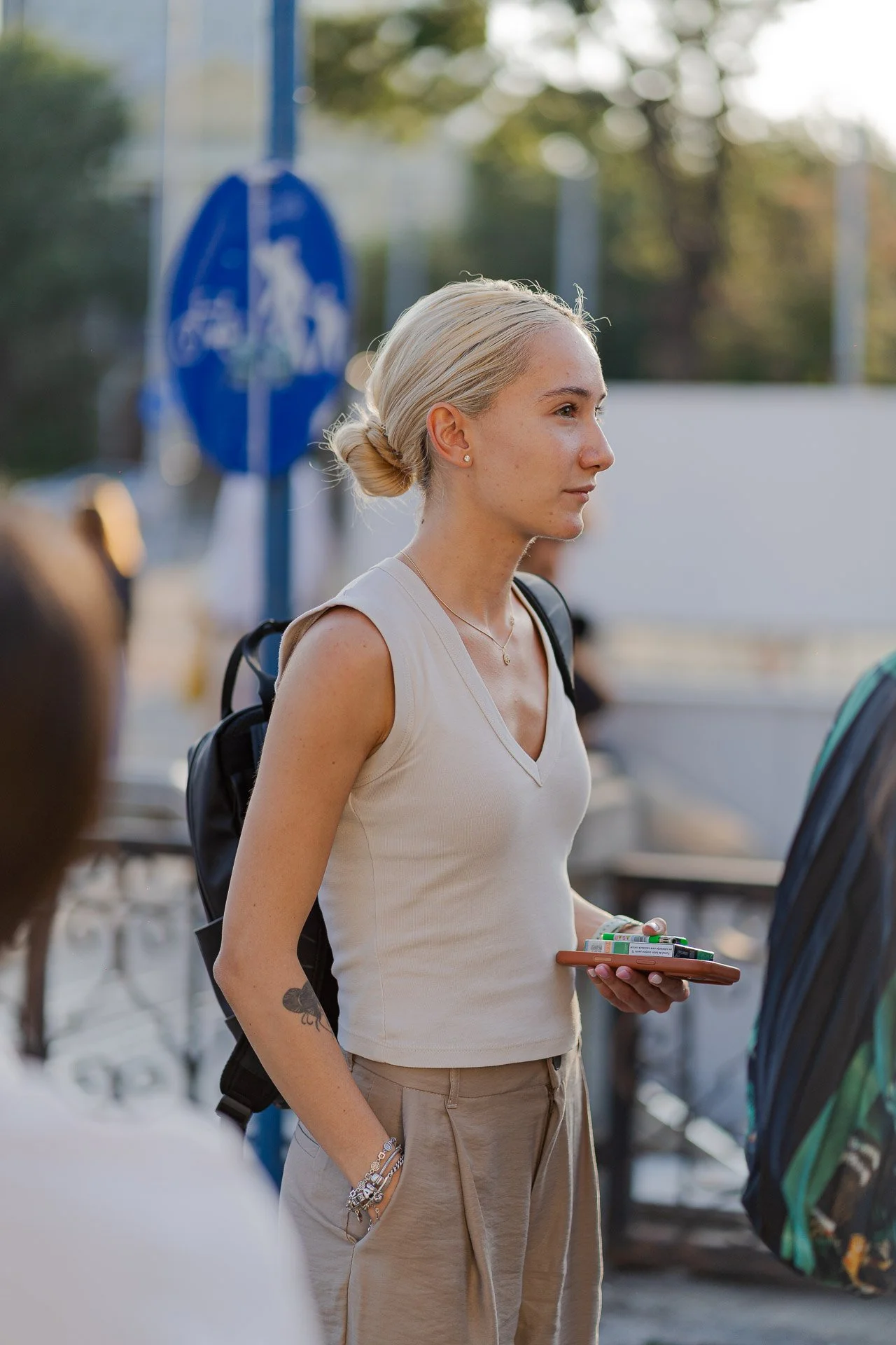 A young woman with blonde hair in a bun, wearing a sleeveless beige top, carrying a backpack, holding a phone with pens and markers, standing outdoors in a crowd.
