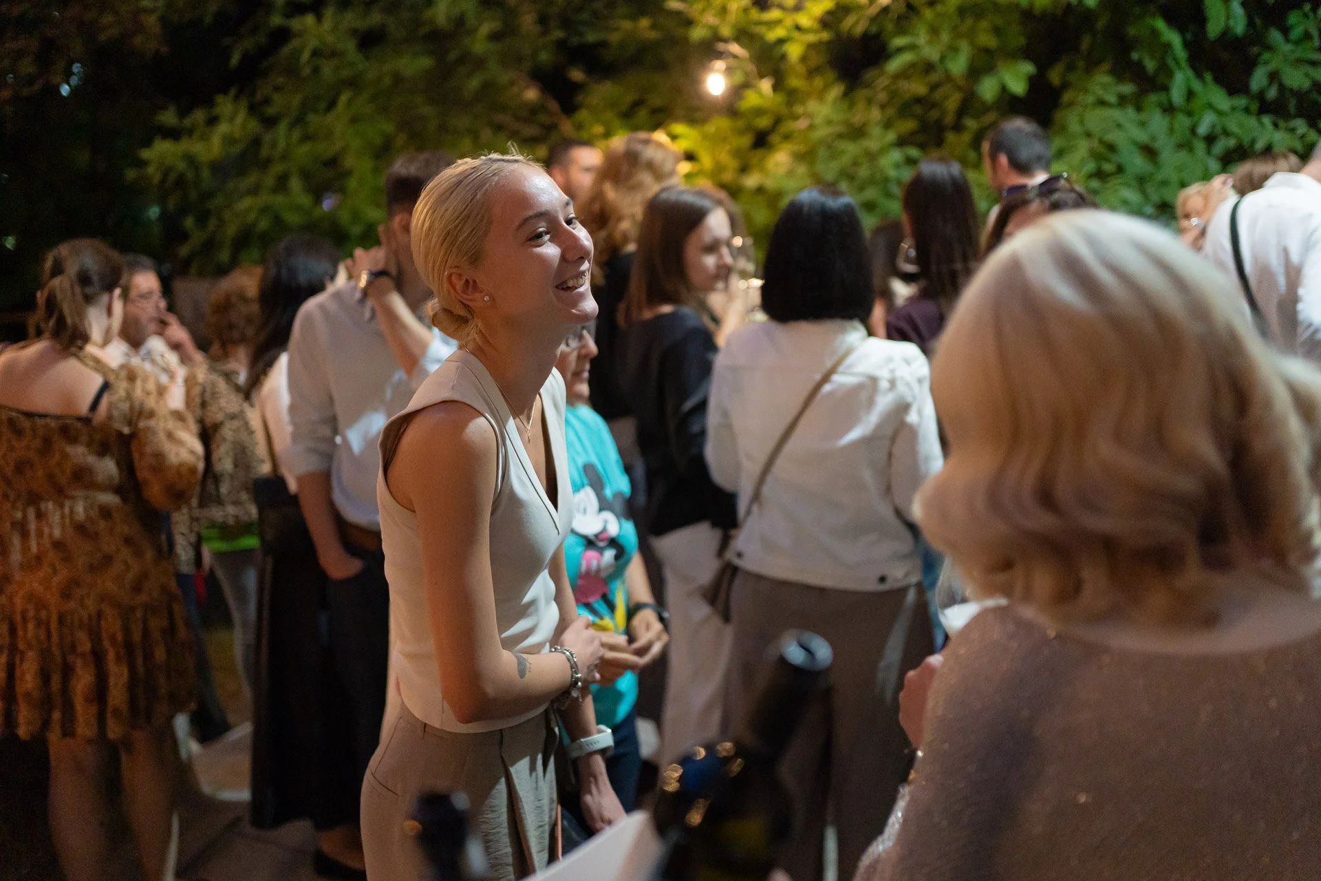 A smiling young woman with blonde hair tied back, wearing a sleeveless beige top, talking to an older woman with blonde hair wearing a sparkly top at an outdoor event in the evening. There are many other people in the background among greenery and di