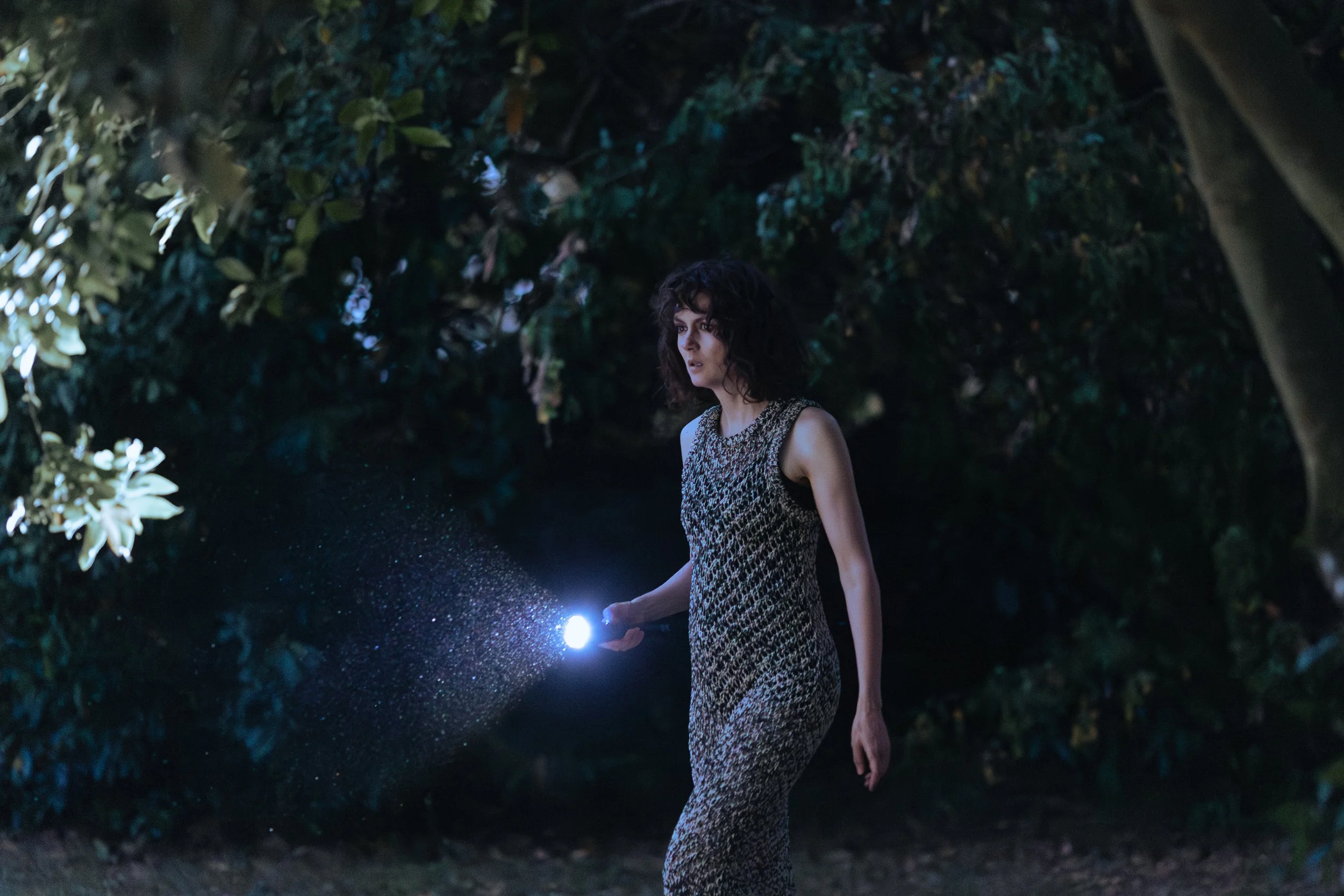 Une femme debout dans l'obscurité, tenant une lanterne qui émet une lumière blanche. Elle porte une robe à motif noir et blanc, et se trouve dans un environnement extérieur avec des arbres en arrière-plan.