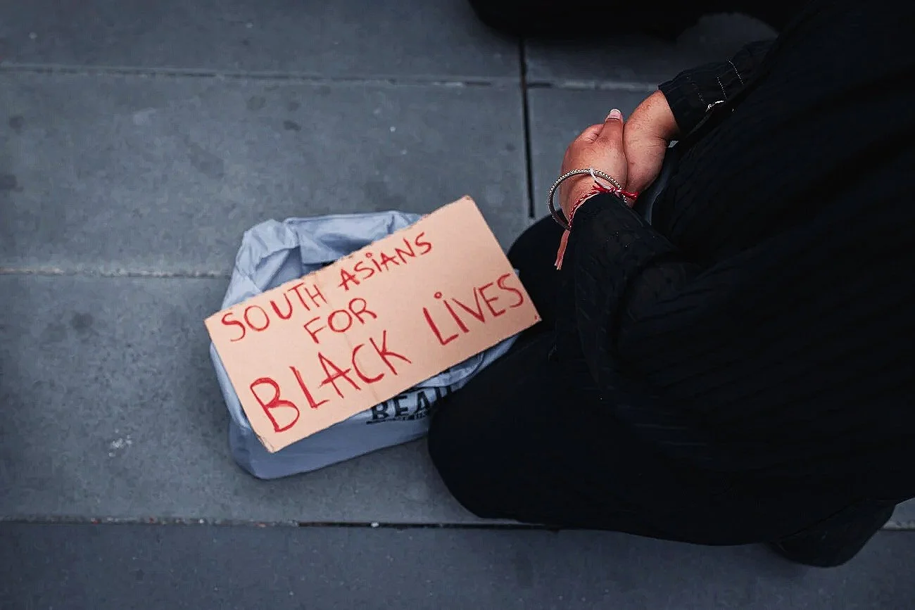 Personne agenouillée avec une pancarte en carton "South Asians for Black Lives" posée sur un sac, sur le sol en béton. Les mains de la personne sont croisées.