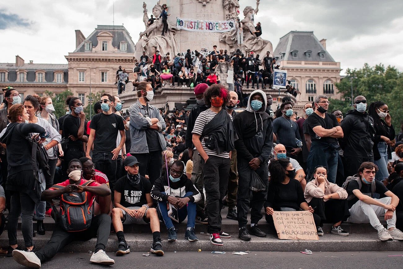 Groupe de personnes portant des masques lors d'une manifestation devant un monument, certains tenant des pancartes avec des messages politiques.