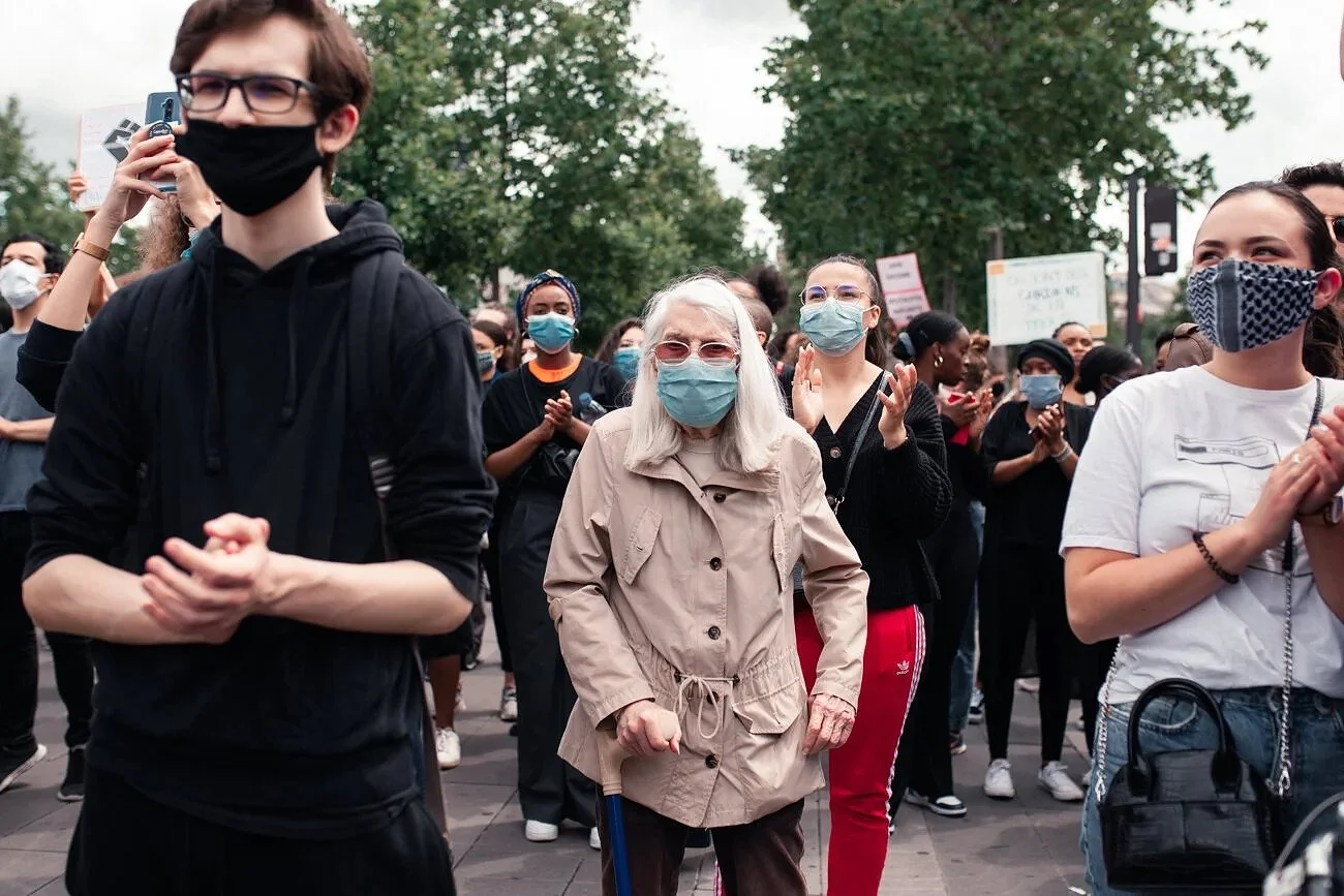 Des personnes portant des masques faciaux lors d'une manifestation en plein air, avec des arbres en arrière-plan.