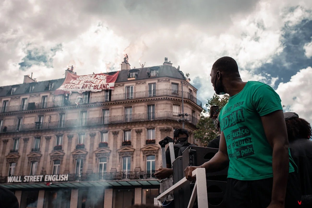 Manifestation à Paris avec des personnes portant des t-shirts verts. Un bâtiment affiche une bannière rouge avec un message. Ciel nuageux.