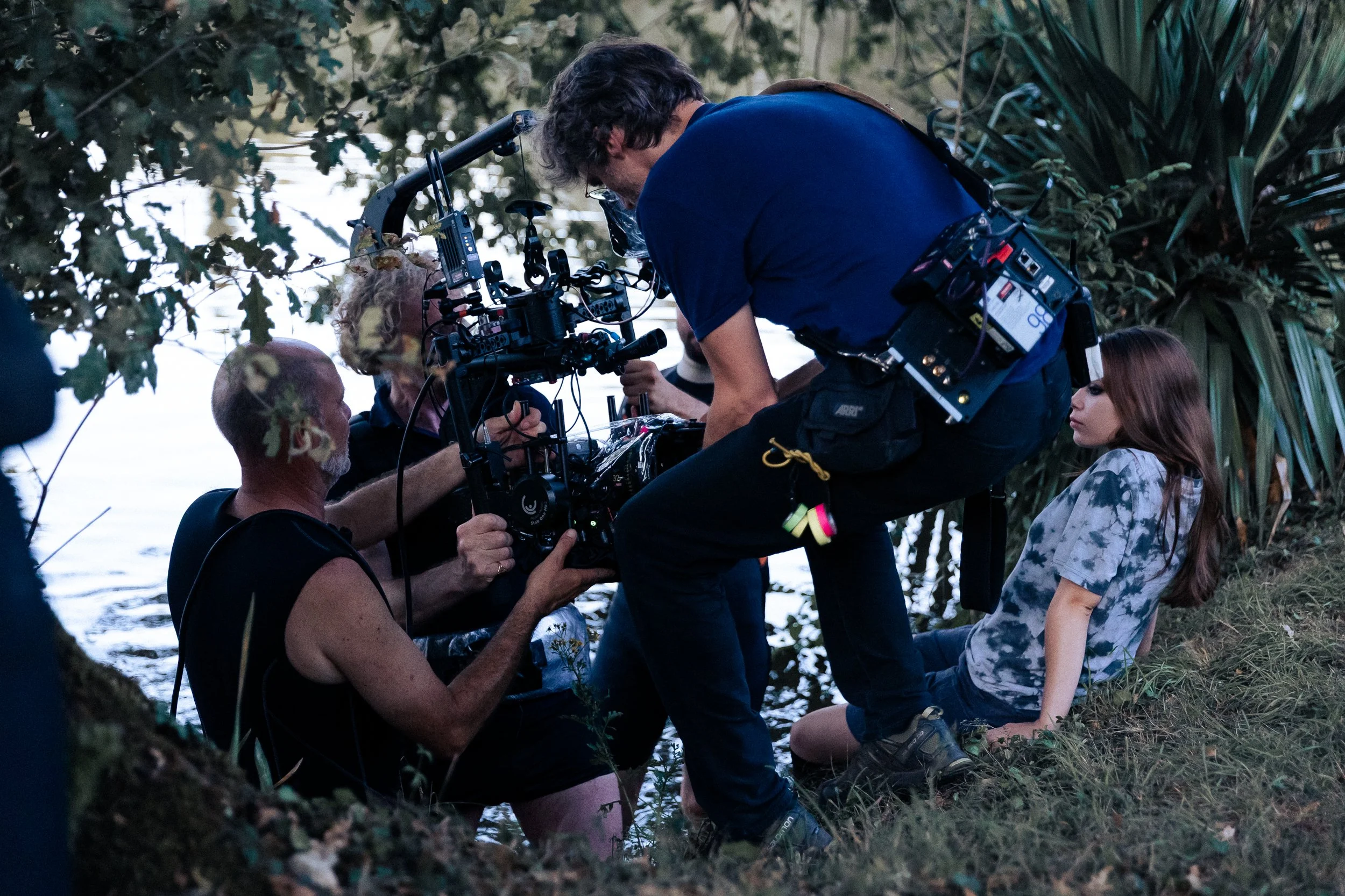 Une équipe de tournage en pleine séance de prise de vue en extérieur, près d'une rivière ou d'un plan d'eau, entourée de végétation dense.