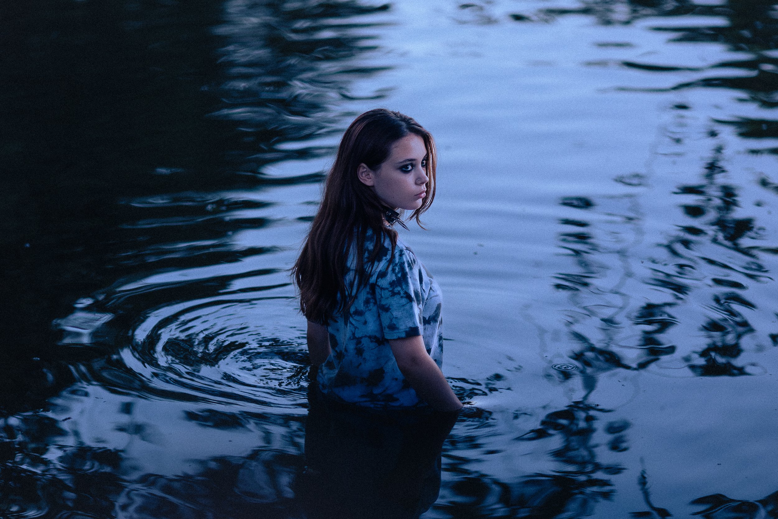 Une jeune femme aux cheveux bruns et vêtue d'un t-shirt à motif finit de se baigner dans un corps d'eau calme, avec un paysage de nature environnante. L'ambiance est sombre et mystérieuse, probablement au crépuscule ou à l'aube.