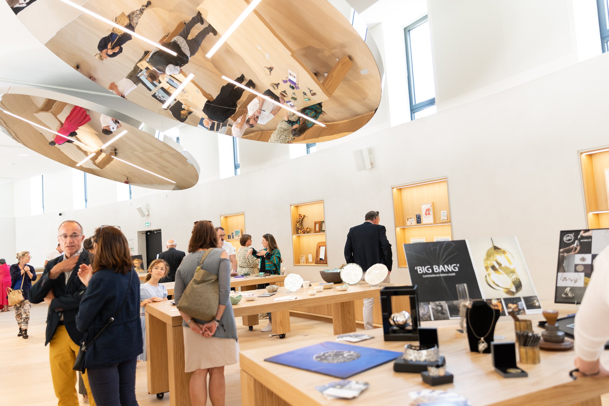 Intérieur d'une galerie moderne avec des vitrines lumineuses et des miroirs réfléchissants au plafond, plusieurs personnes regardent des objets exposés sur des tables en bois.