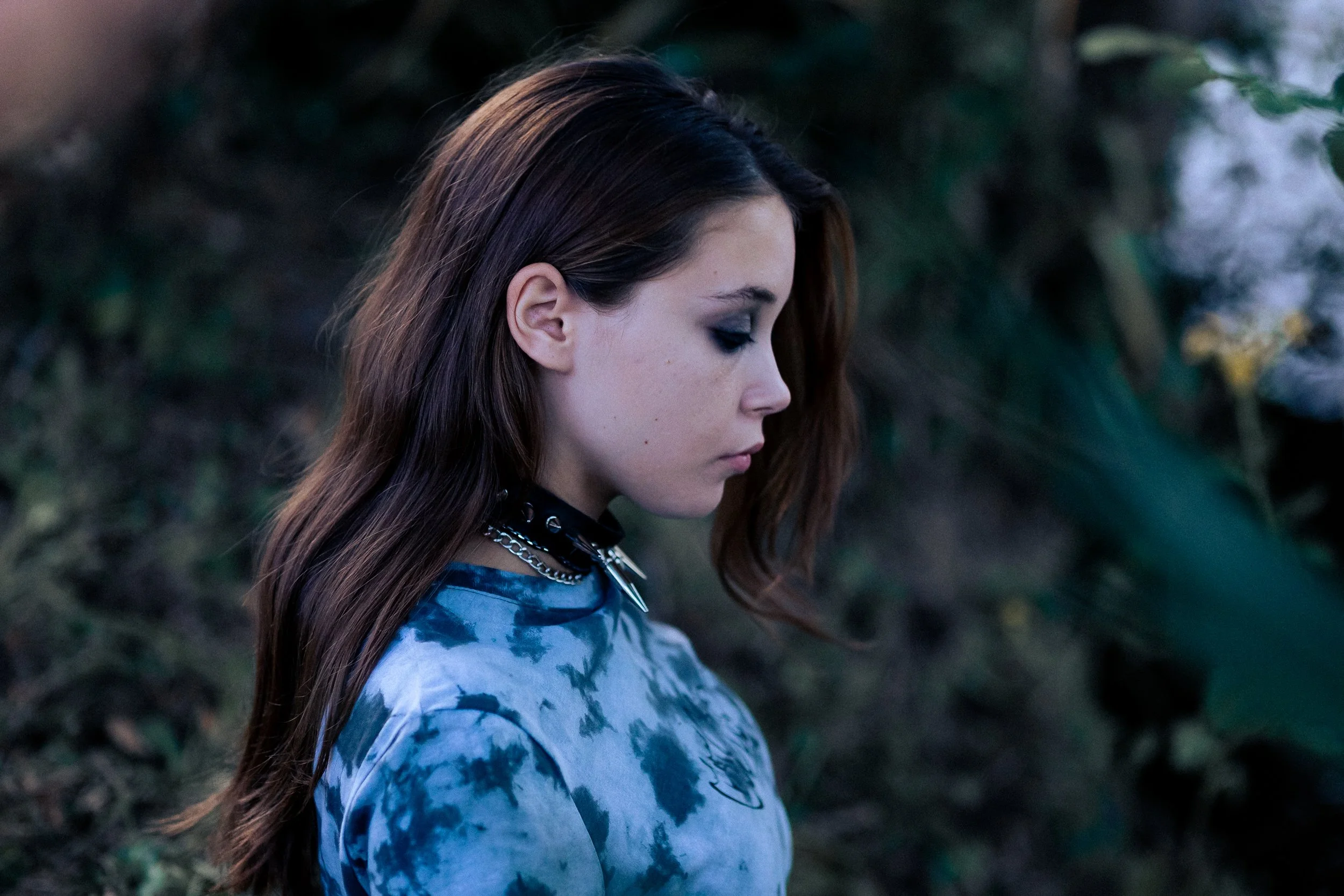 Jeune femme aux cheveux longs, bruns, portant un maquillage sombre et un collier en chaîne, regardant vers le bas dans un environnement naturel.