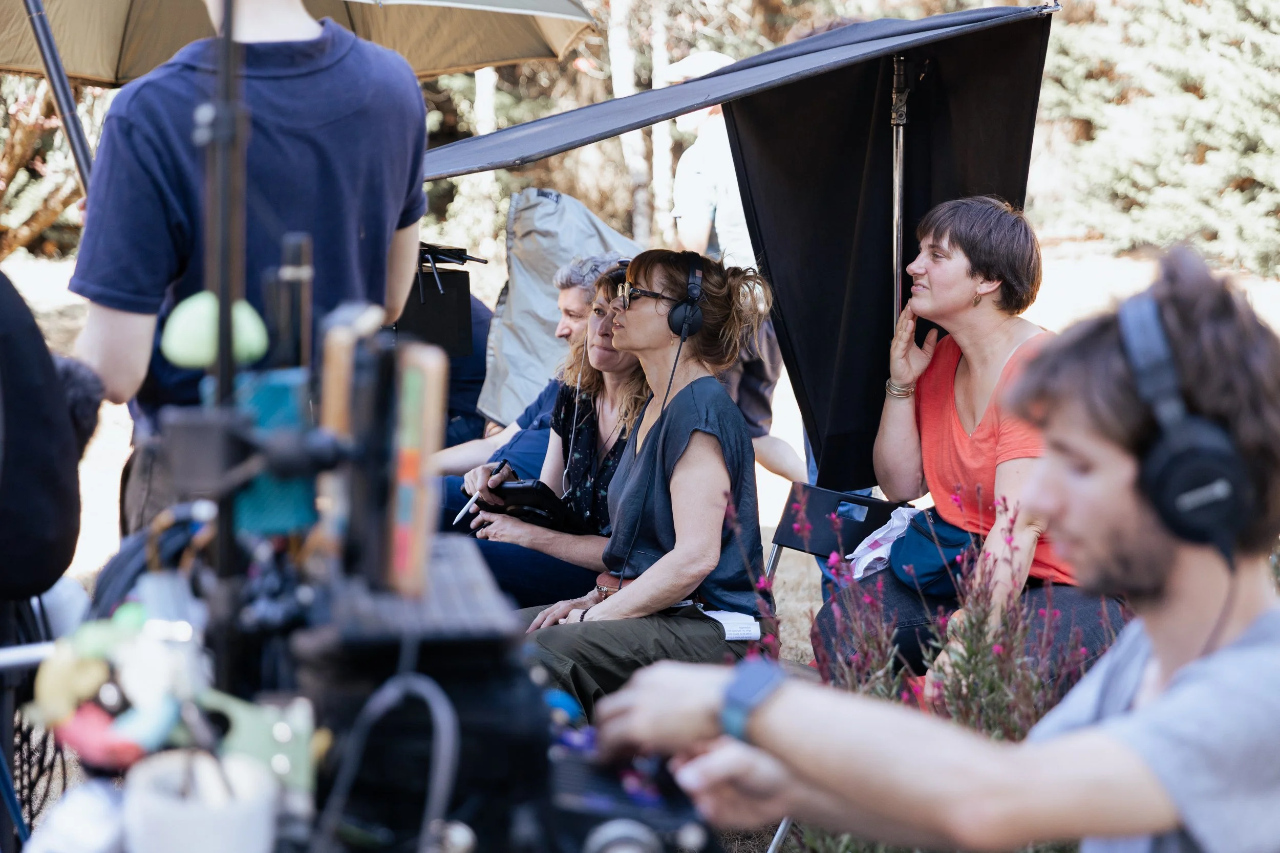Une équipe de tournage en plein extérieur, avec plusieurs personnes assises sous un abri, dont une porte un casque audio, alors qu'un autre préparant le matériel. L'ambiance semble détendue et concentrée, dans un environnement naturel avec des arbres