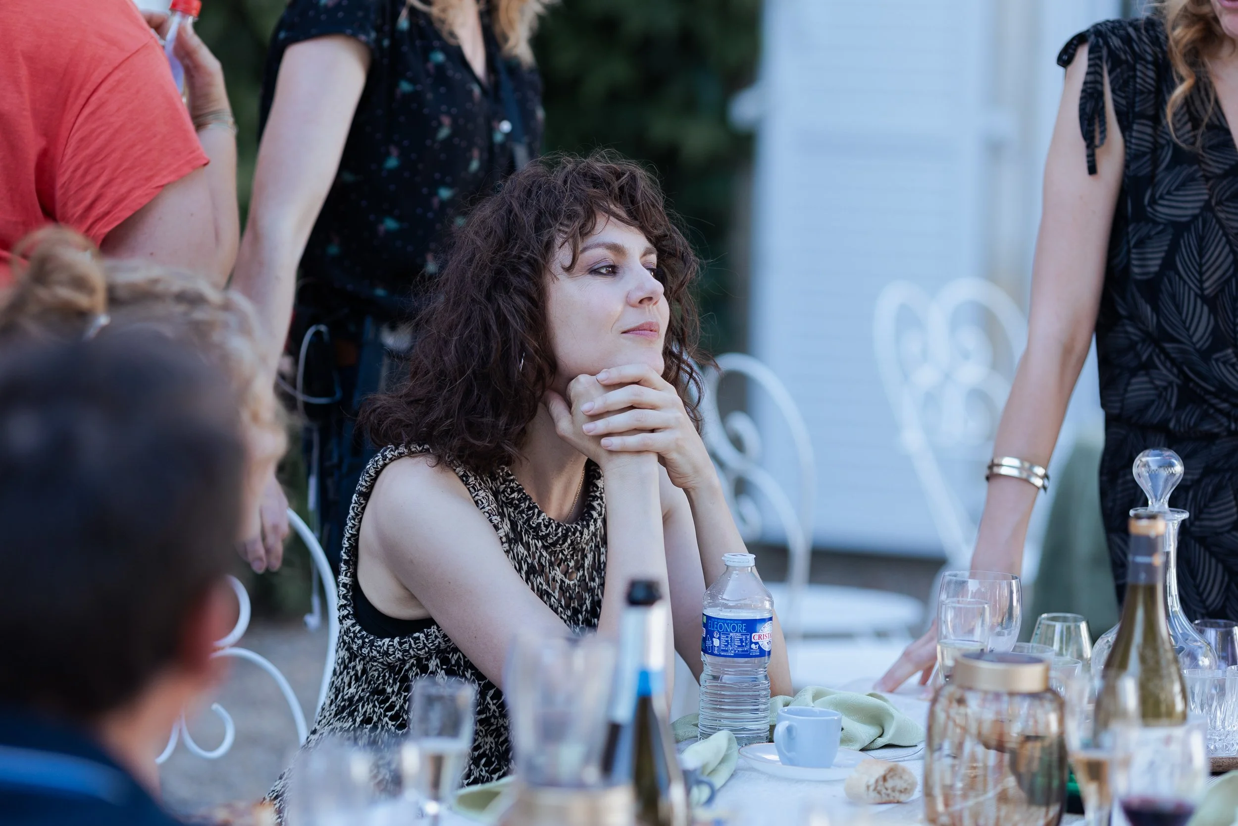 Une femme aux cheveux bouclés et vêtements à motifs assise à une table lors d'un repas ou d'une réunion en extérieur, entourée d'autres personnes.