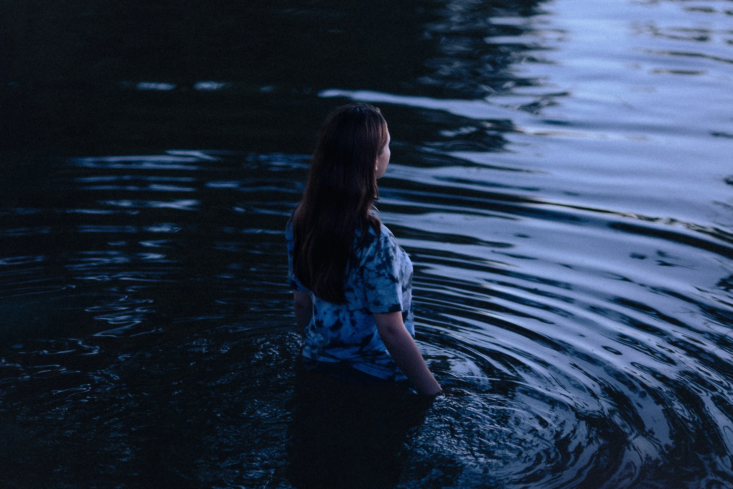 Jeune femme assise dans un étang ou une rivière lors du crépuscule, regardant vers l'eau avec une lumière douce sur la scène.