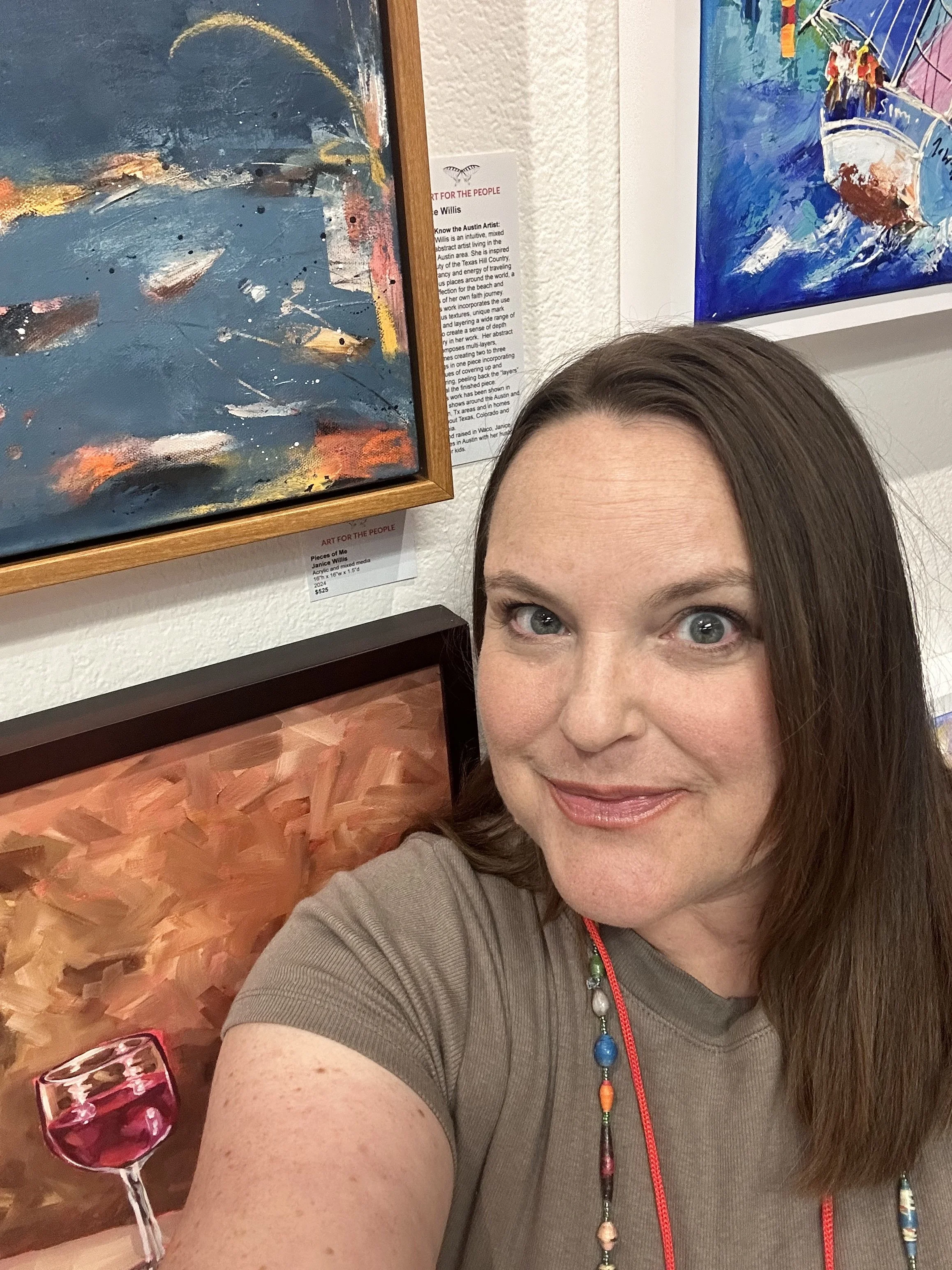 A woman with long brown hair and blue eyes smiling for a selfie at an art gallery. Behind her are several colorful paintings, including an abstract piece with blue, orange, and pink tones, and another painting of a boat on water with vibrant colors.