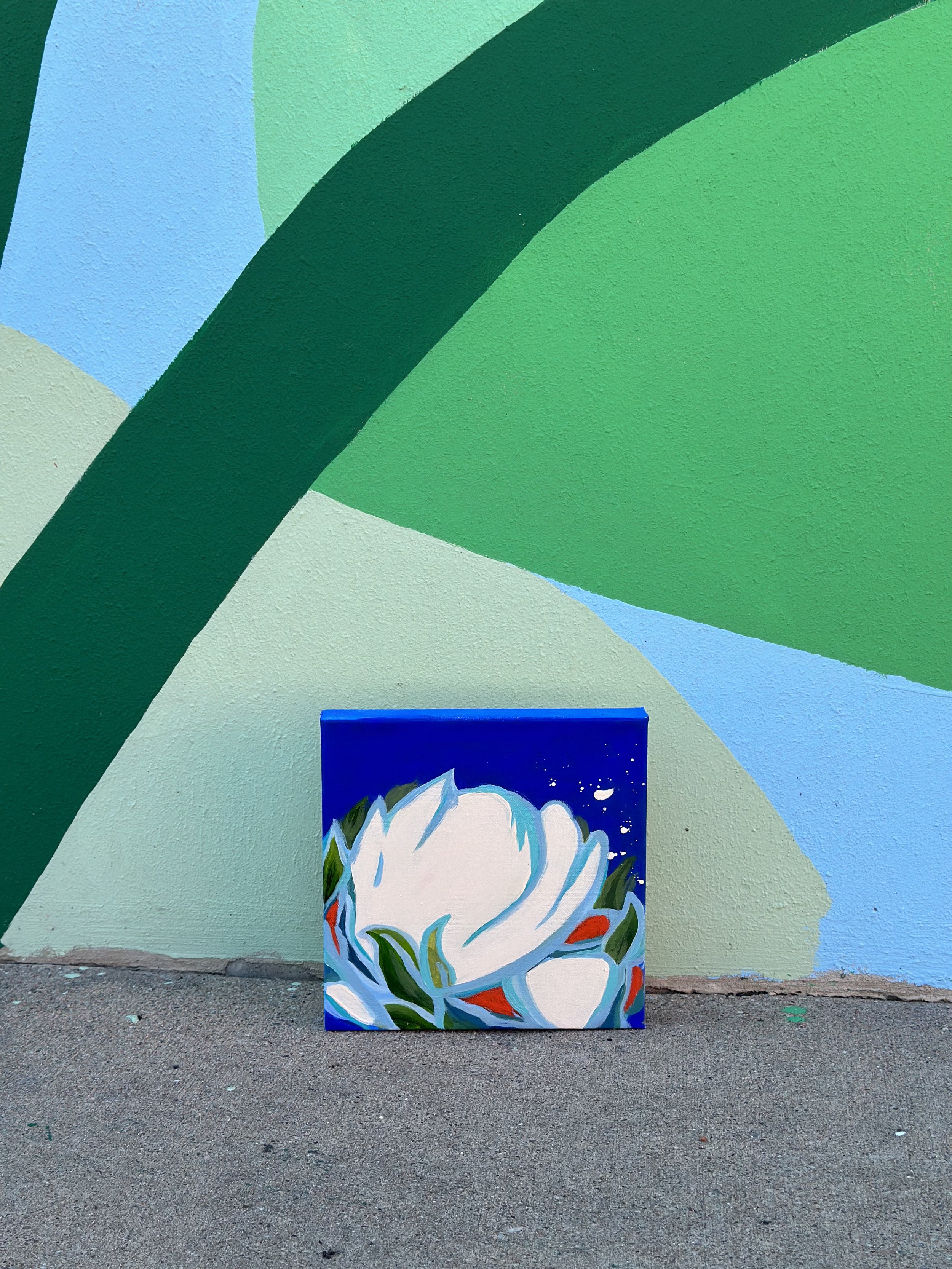 A colorful mural with large green, blue, and light green shapes on a wall, with a small painting of a white flower with green and red accents resting on the ground in front of the mural.