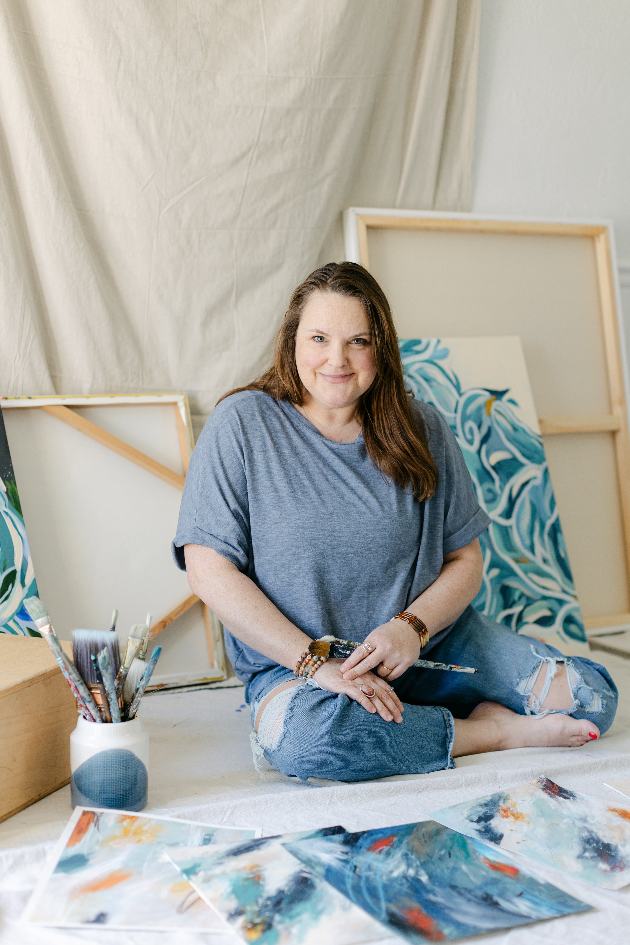 A woman with long brown hair sitting cross-legged on a white cloth, holding a paintbrush, surrounded by completed abstract landscape paintings and painting supplies, with blank canvases behind her.