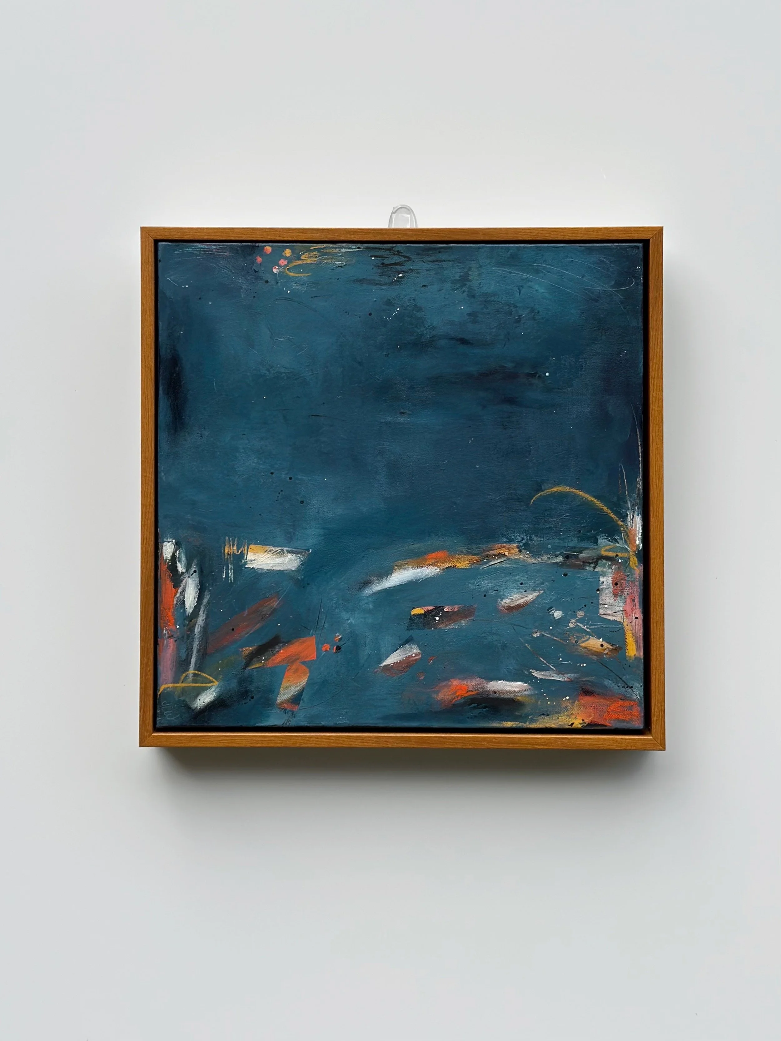 Abstract dark blue painting with splashes of white, orange, and hints of red, framed in wood, mounted on white wall.