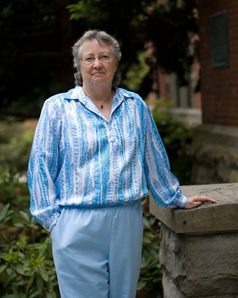 A photo of MaryJane Nordgren with short gray hair and glasses standing outdoors, wearing a blue and white patterned blouse and light blue pants, next to a stone ledge with her hand resting on it.