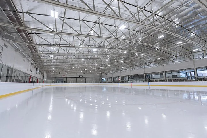 Empty indoor ice skating rink with bright lighting and a high arched ceiling.