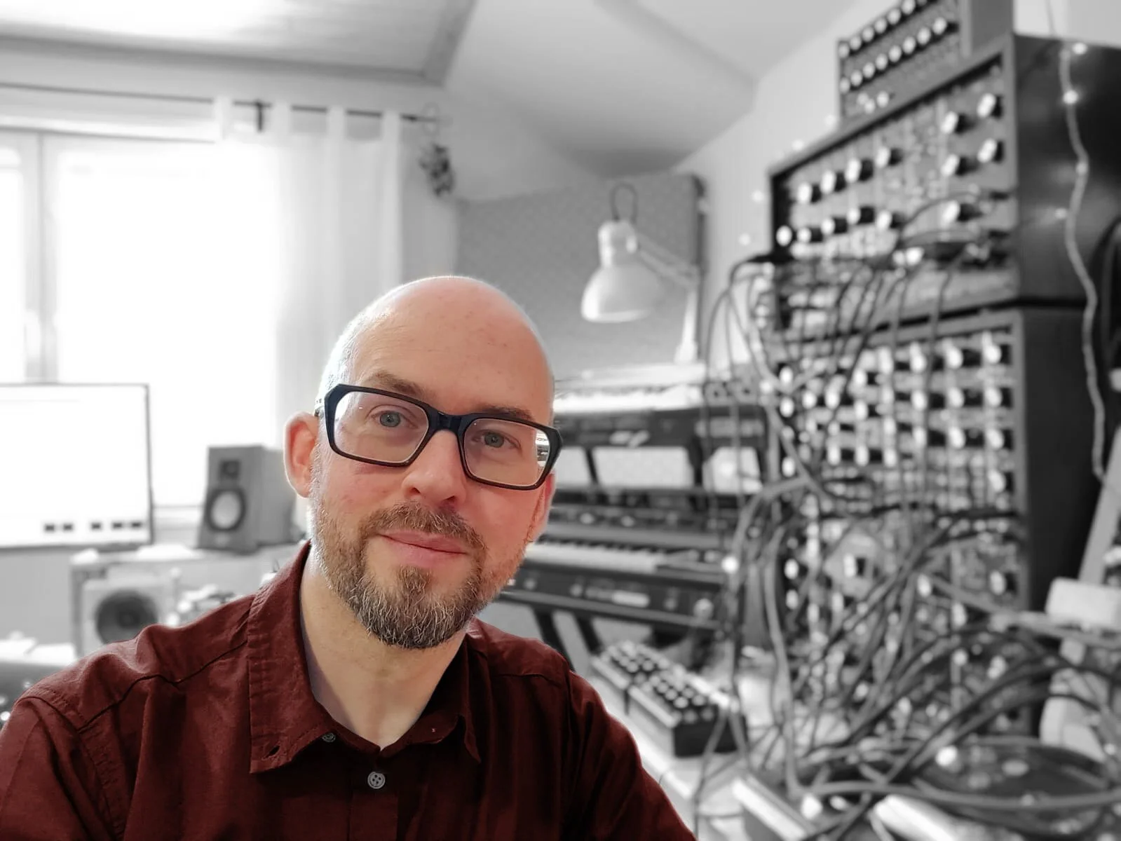 A man with glasses and a beard taking a selfie in a room filled with electronic music equipment, including synthesizers, cables, and a computer monitor, with a window in the background.