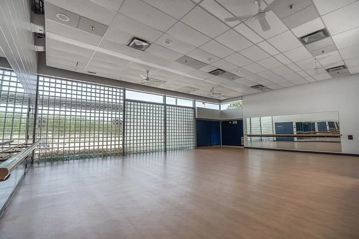 An empty dance or exercise studio with large windows, a wooden floor, mirrors, and ceiling fans.